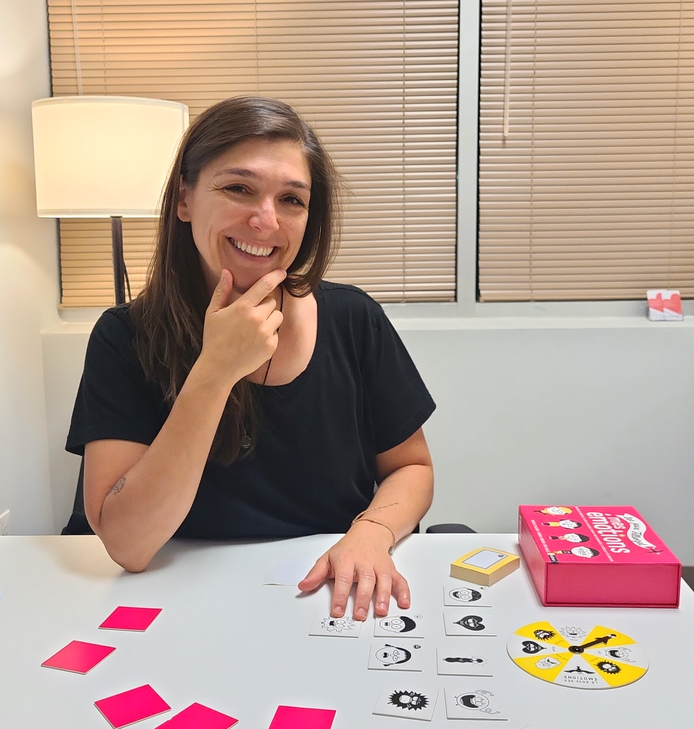 Une femme joue à un jeu de société sur une table blanche. Elle a les cheveux bruns, porte un haut noir et sourit. Sur la table, il y a des cartes, des jetons, et un outil de jeu. En fond, il y a des stores en bois et une lampe de bureau.