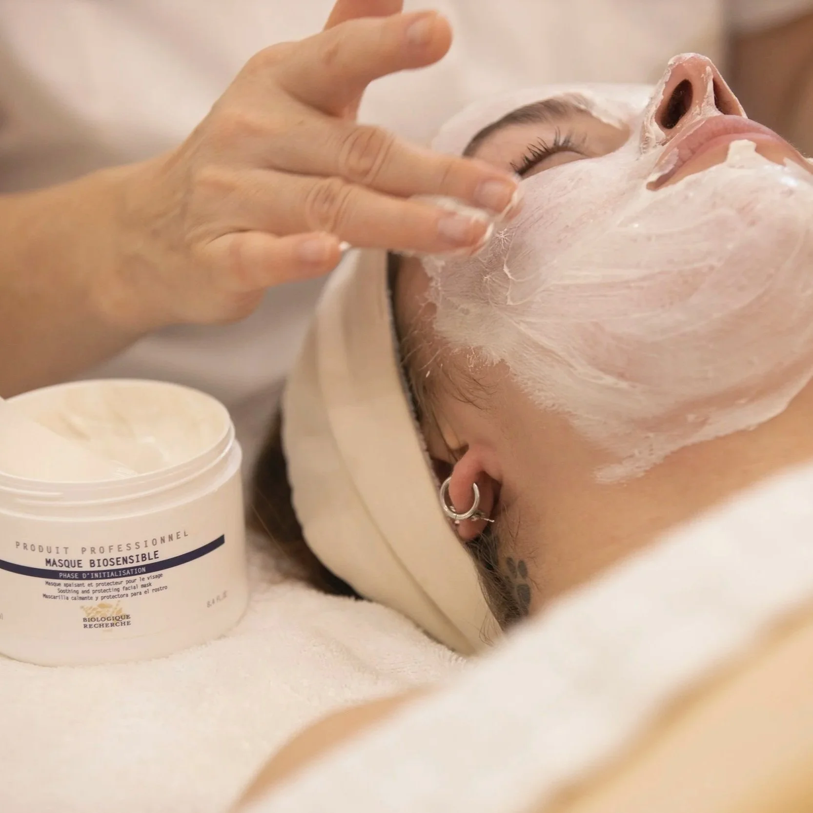 A woman receiving a facial massage with a thick white facial mask applied to her face. The therapist is gently touching her face, and the woman is lying down with closed eyes inside a spa or skincare setting.