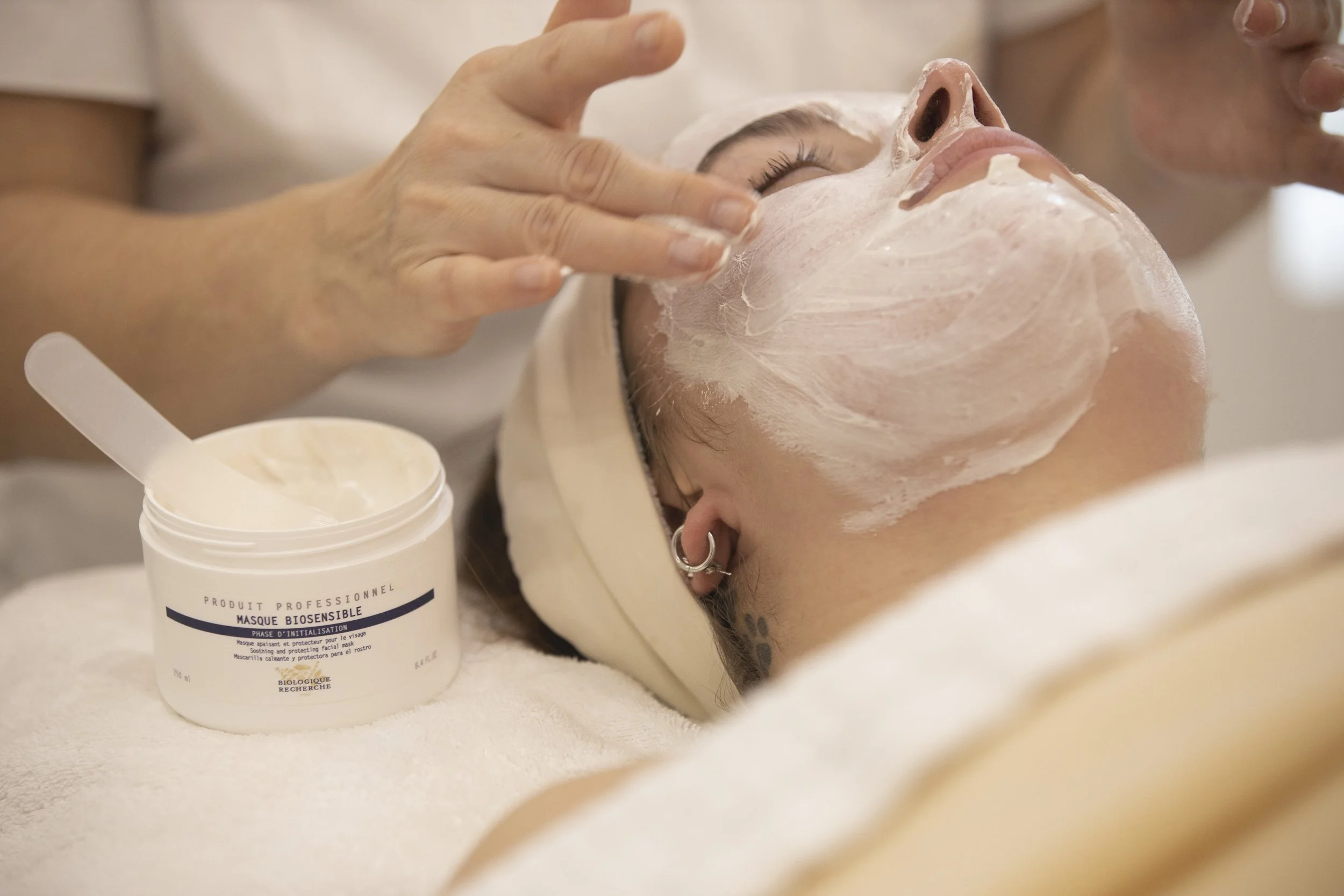 A person receiving a facial treatment with a facial mask applied on their face, while a salon professional tends to them, with a container of facial mask nearby.