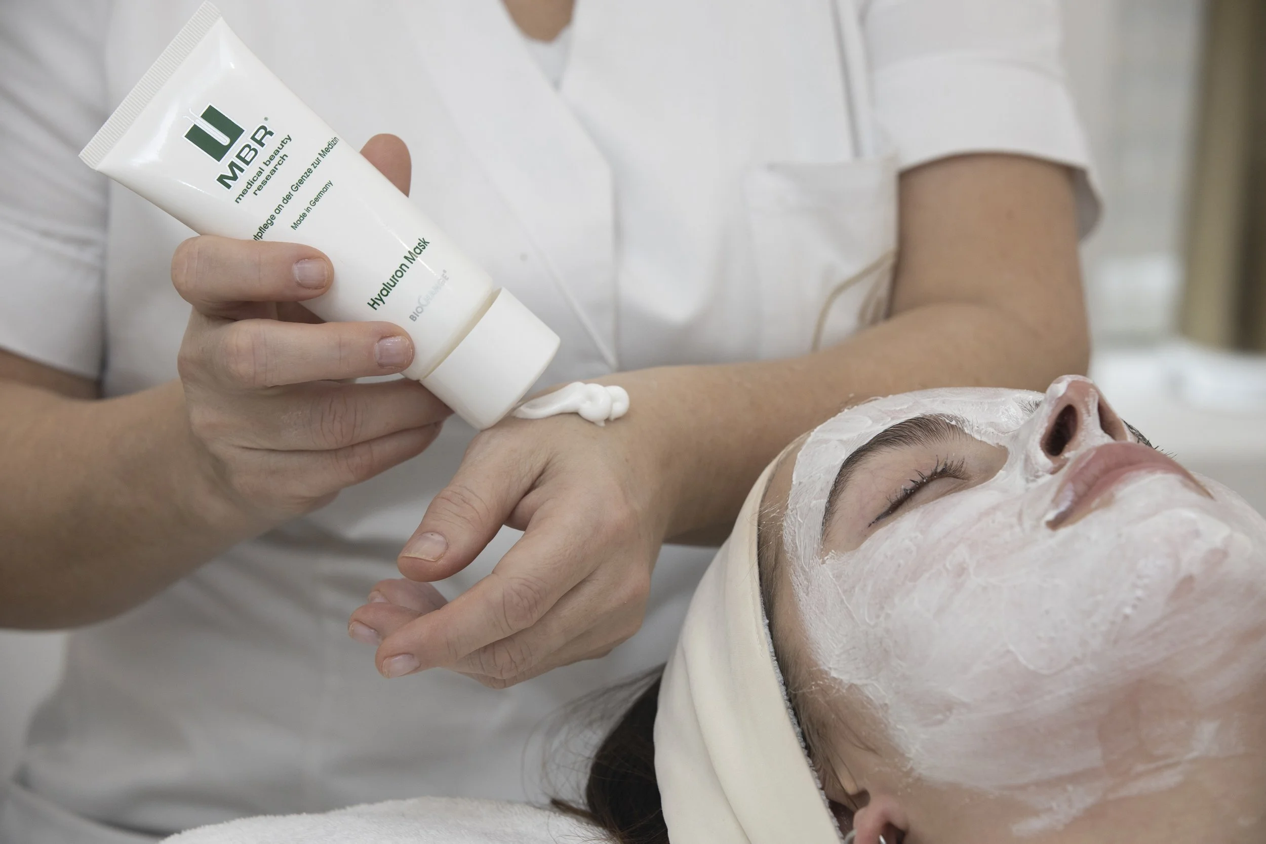 A person receiving a facial treatment at a spa, with a practitioner applying a white cream mask to their face, and the person lying on a treatment bed with their eyes closed