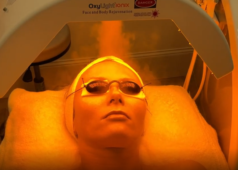 Person lying under a UV light for facial therapy, with protective goggles on, in a medical or cosmetic treatment room.