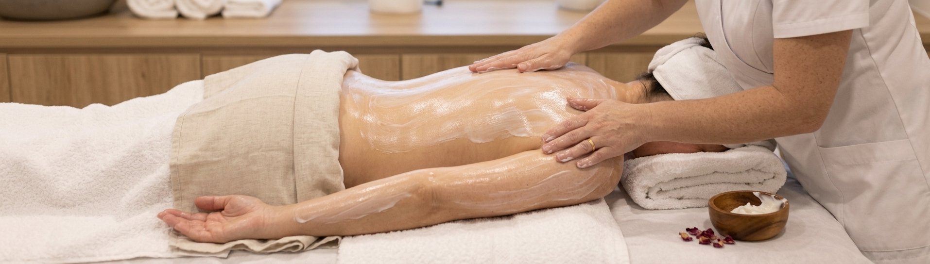 A person receives a massage on their back in a spa or massage room, lying face down on a massage table with towels underneath and a cream or lotion applied to their back.
