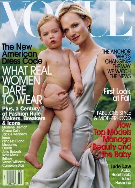 Vogue magazine cover featuring a woman holding a young child, both dressed minimally, with a scenic outdoor background and magazine headlines.