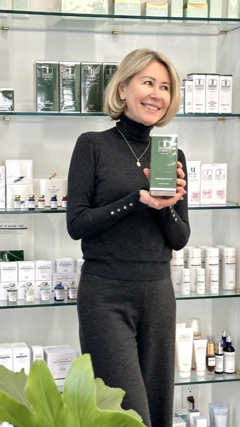 A woman with blonde hair wearing a black turtleneck and pants, smiling while holding a green skincare product box in a store with glass shelves filled with skincare products behind her.