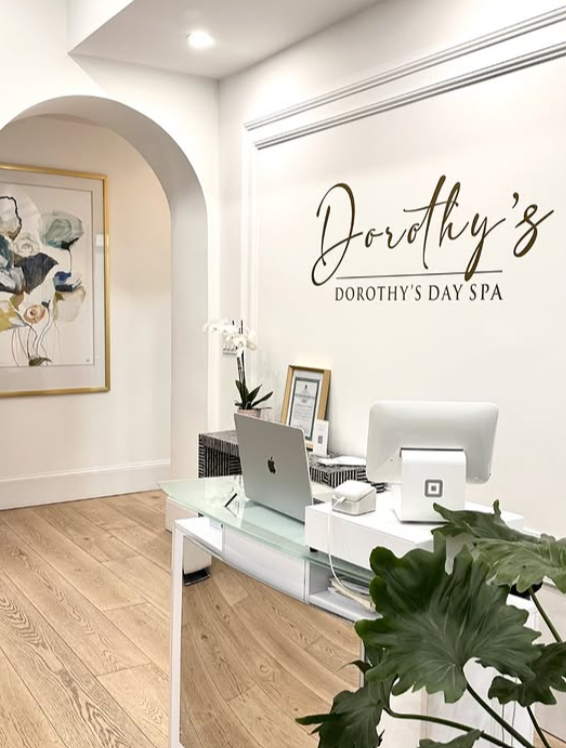 Reception area at Dorothy's Day Spa with a white desk, iMac computer, plant, framed certificate, and wall sign reading 'Dorothy's' in cursive with smaller print underneath.