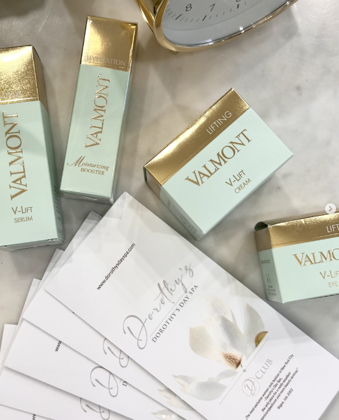 Several Valmont skincare products, including V-Lift Serum, Moisturizing Booster, V-Lift Cream, and Eye Cream, are arranged on a marble surface, along with some informational pamphlets from Dorothy's Day Spa.
