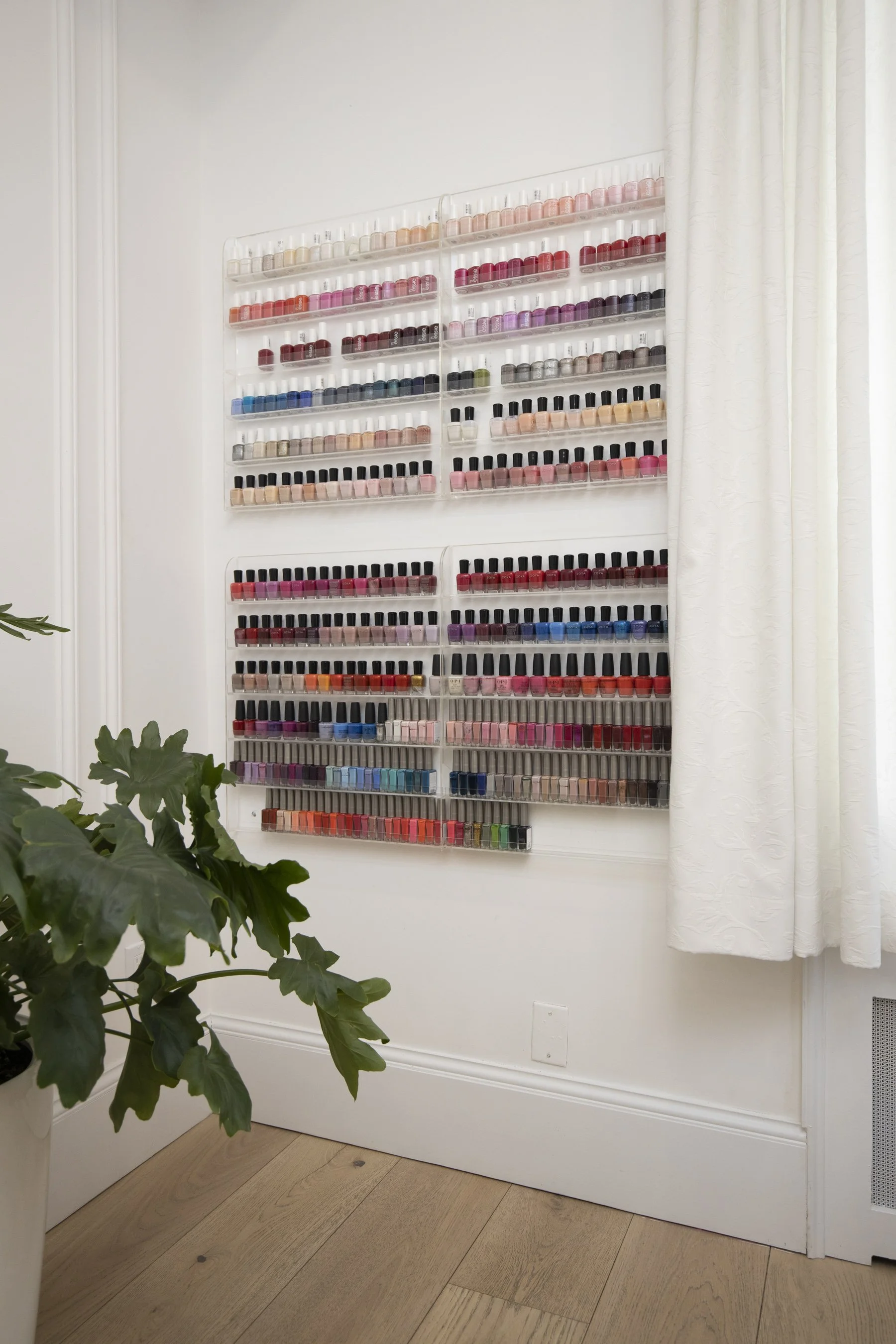 A wall-mounted display of multiple rows of colorful nail polish bottles in a bright, white room with hardwood flooring and a white curtain on the right side. A large green plant is in the lower left corner.