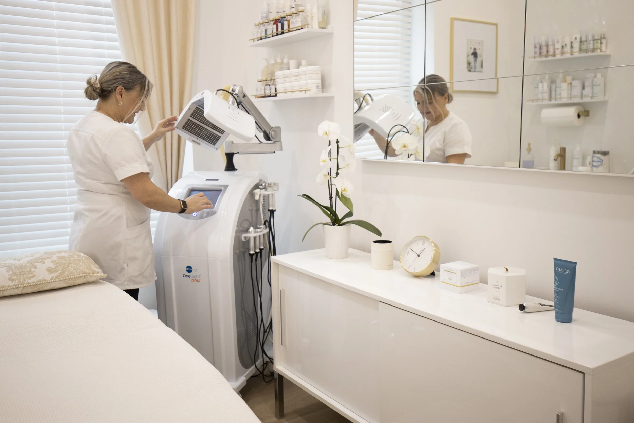 A woman in a white uniform is operating a medical or cosmetic device in a well-lit clinic room. The room has white walls, a large mirror, shelves with bottles, a potted orchid, and a bedside table with skincare products and a clock visible.
