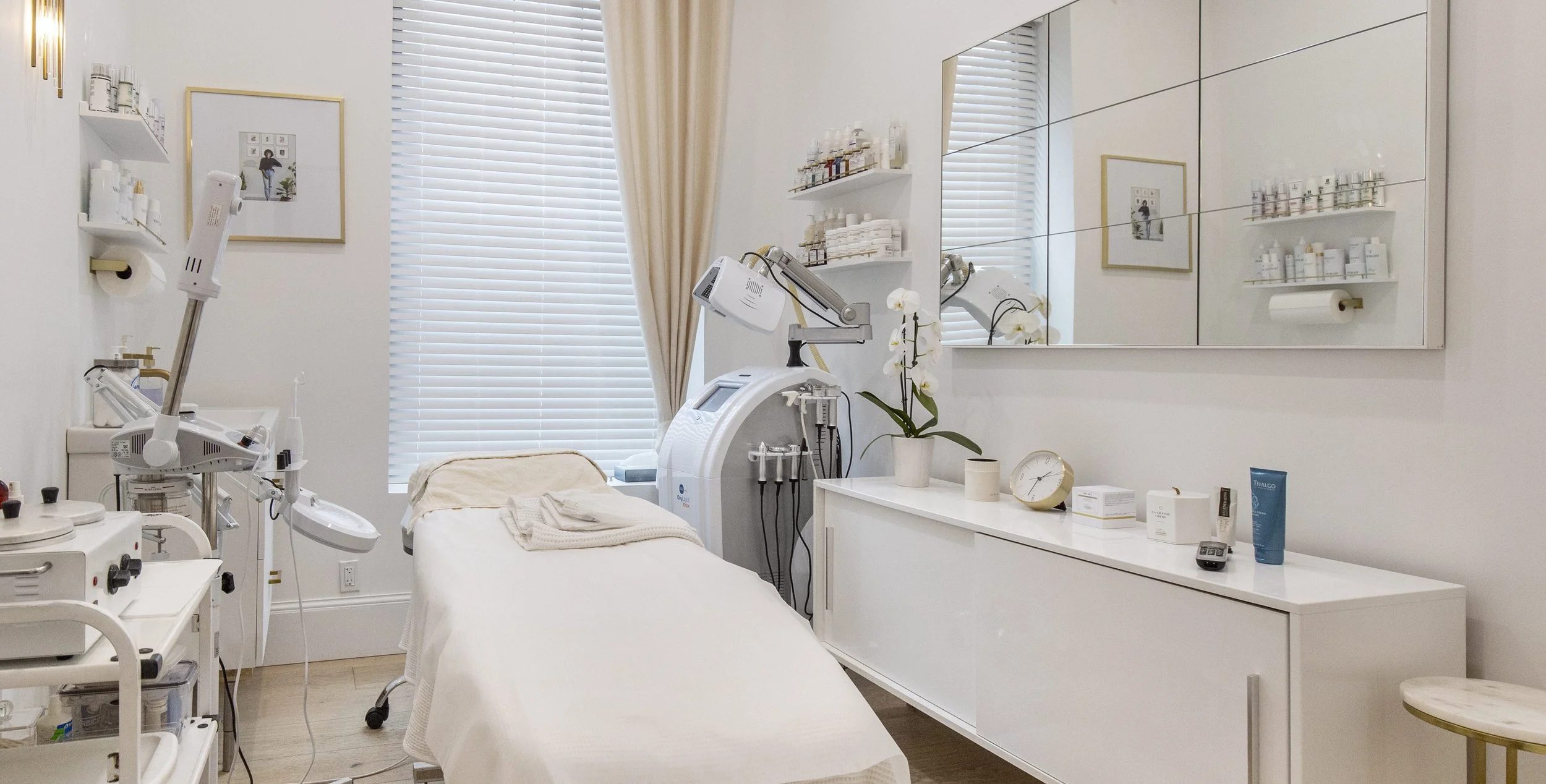 A treatment room in a medical or cosmetic clinic with a treatment bed, medical equipment, shelves with products, a large mirror, and a window with blinds.