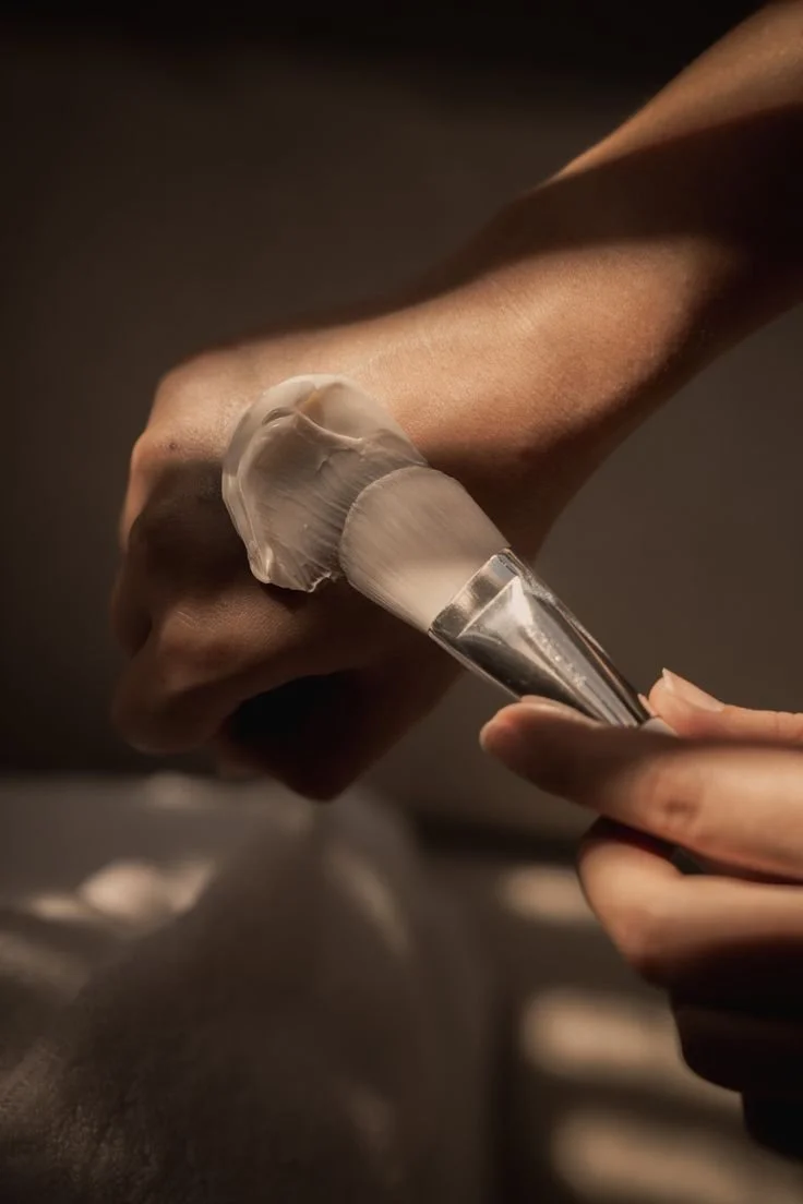 Person applying face cream or moisturizer with a brush on their cheek.
