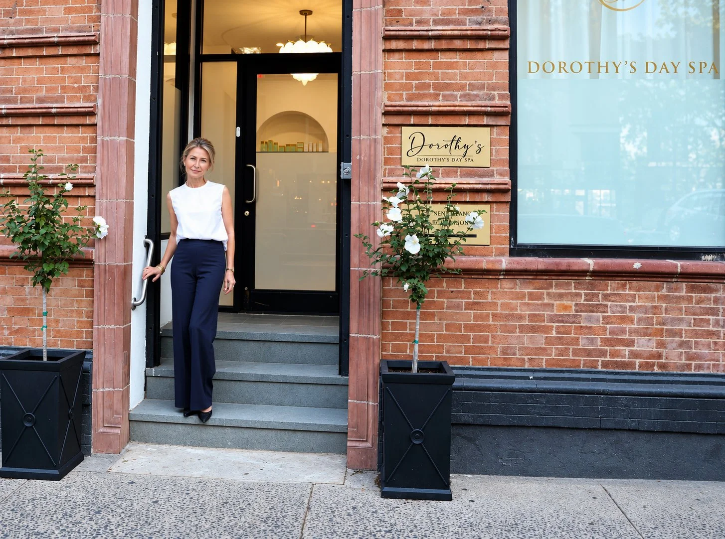 A woman standing on the steps outside Dorothy's Day Spa in a sleeveless white top and navy blue pants, with potted plants on either side of the entrance.