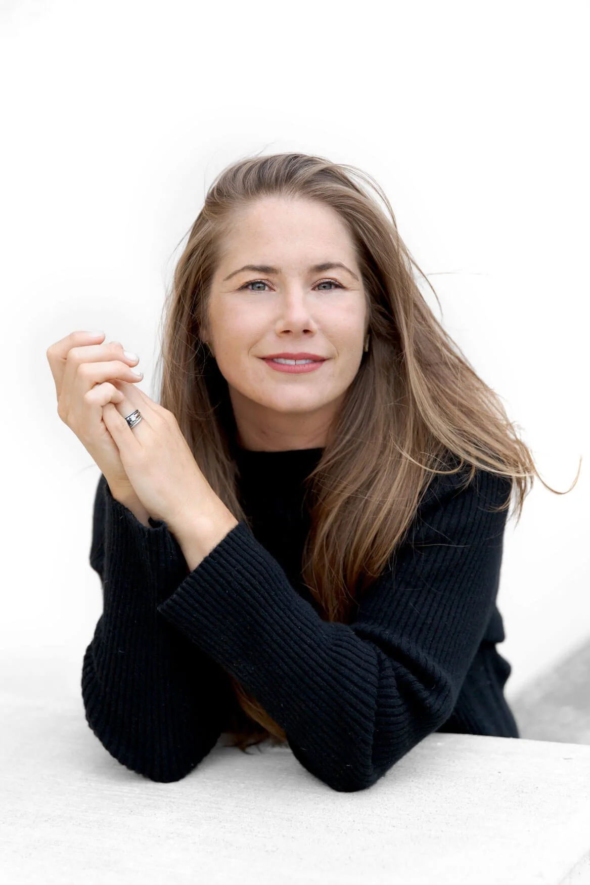 A woman with long, light brown hair is smiling and looking at the camera. She is resting her chin on her hands, which are clasped together, and wearing a black, ribbed long-sleeve shirt with rings on her fingers. She is sitting against a plain white background.