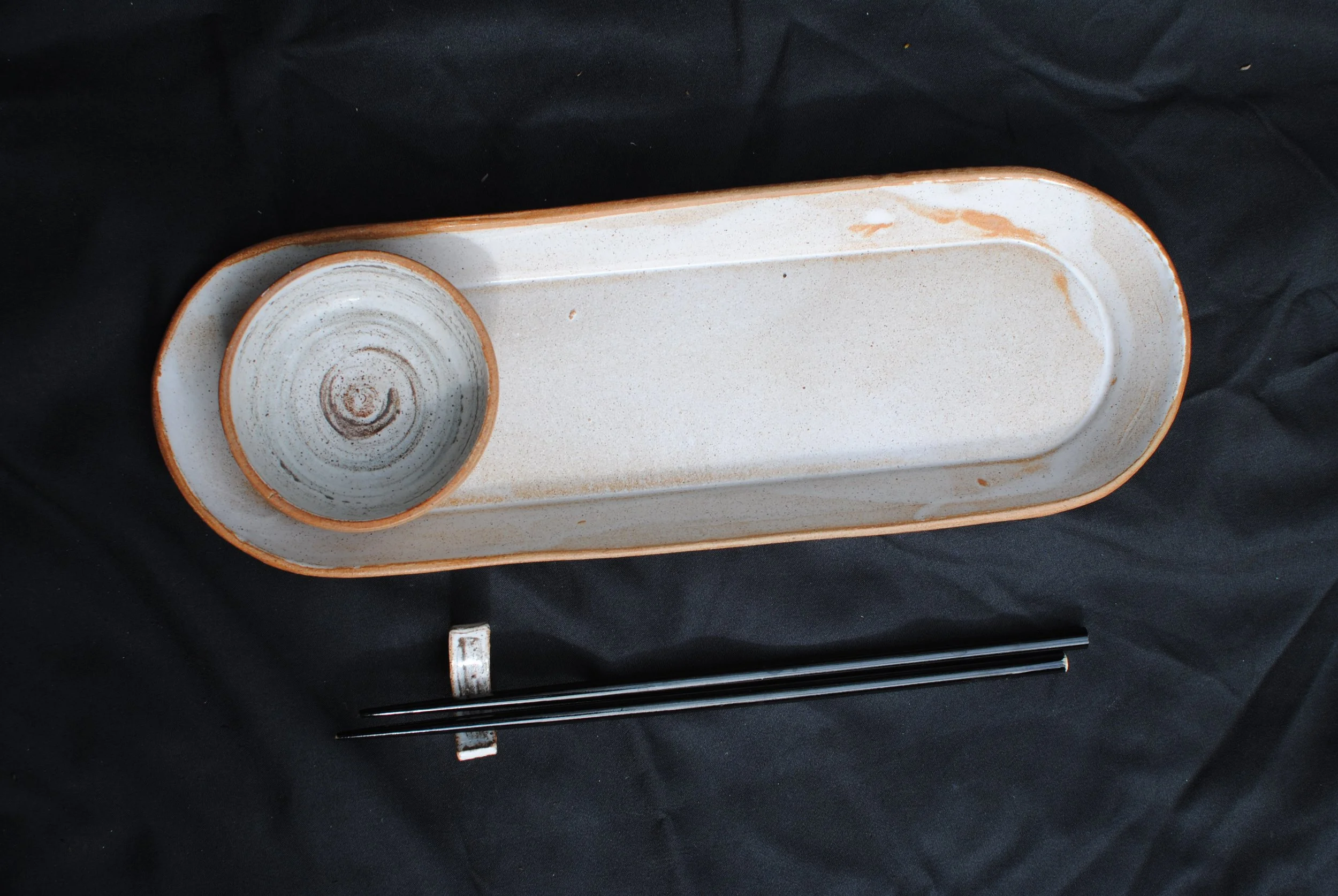 A rectangular ceramic tray with a small circular bowl at one end, both featuring a speckled beige and brown glaze, resting on a black fabric background, with a black chopstick placed underneath the tray.