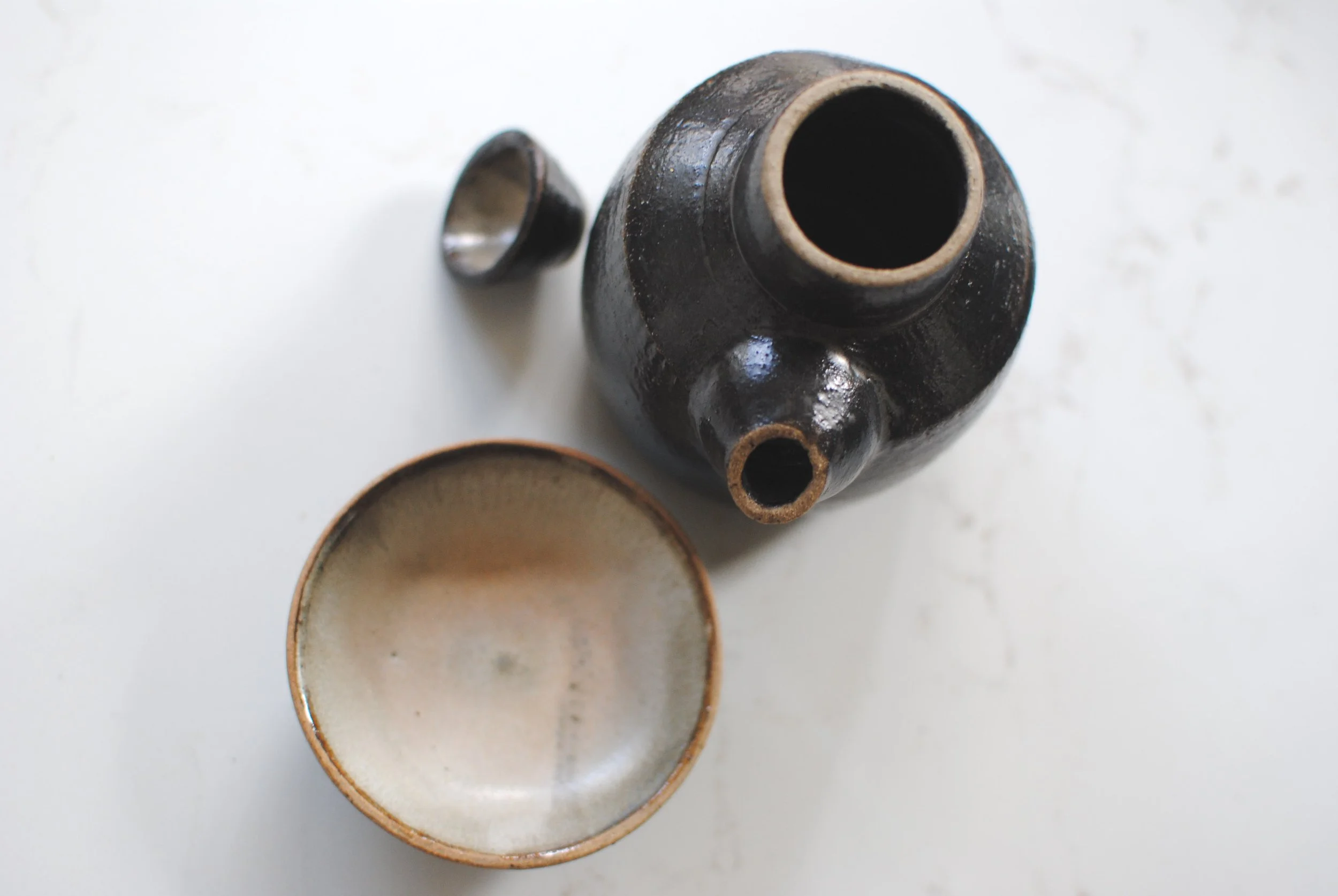 A black ceramic teapot with a round spout and lid, viewed from above, with a white ceramic cup beside it on a white surface.