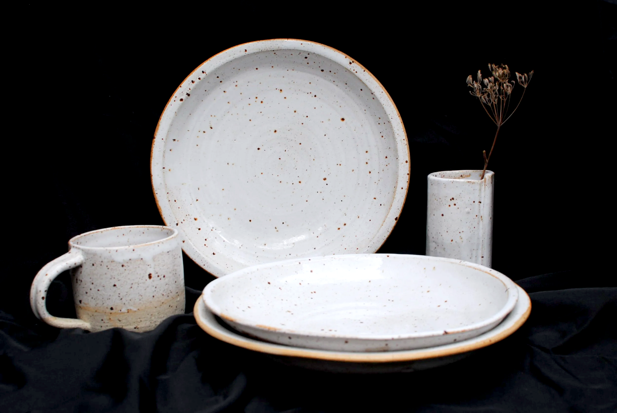 A collection of white ceramic dishes, including a large plate, a mug, and small bowls, with a dried flower in a small vase, all set against a black background.