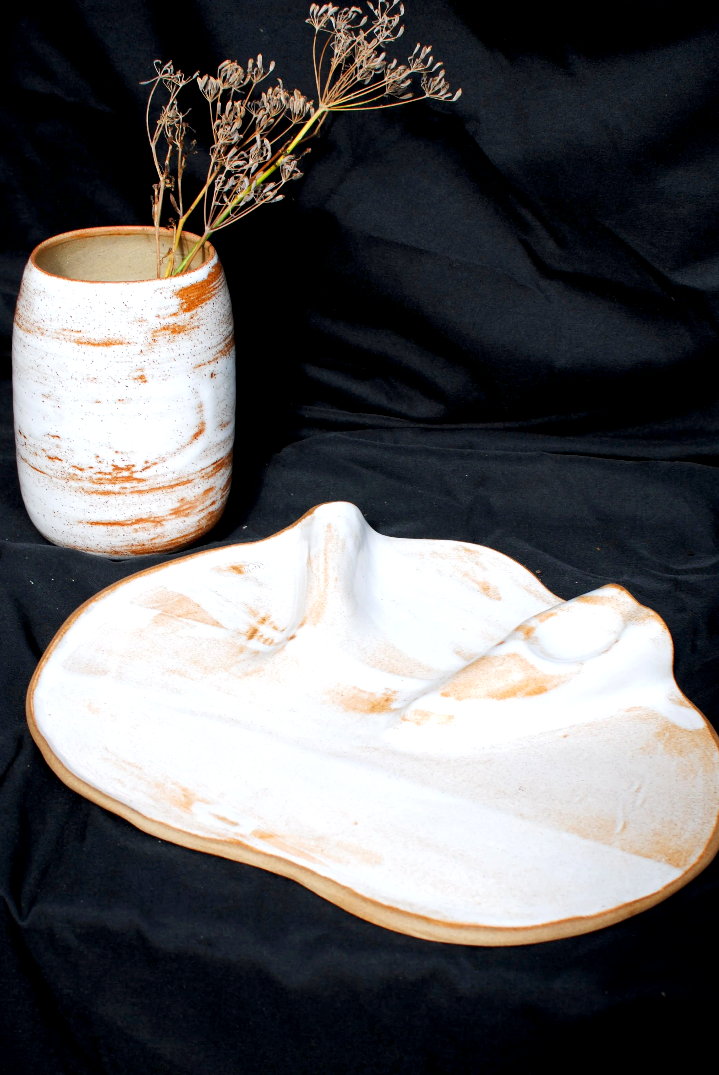 A ceramic vase with a brown and white marbled pattern holding dried plants, placed on a black fabric background, with part of a white ceramic dish in the foreground.