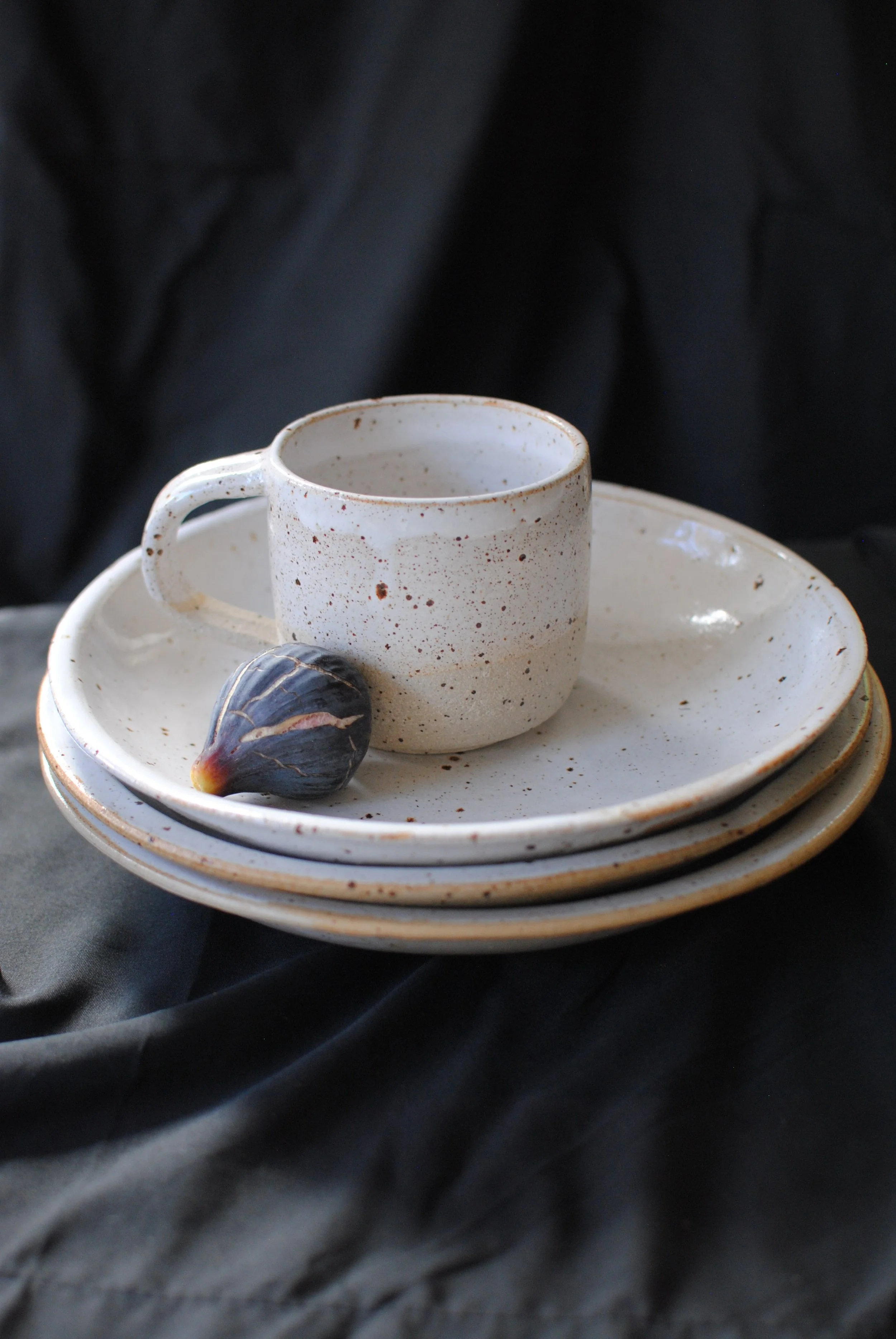 A white ceramic cup with brown speckles on a matching saucer, and a single dark fig with purple and yellow hues, set against black fabric background.