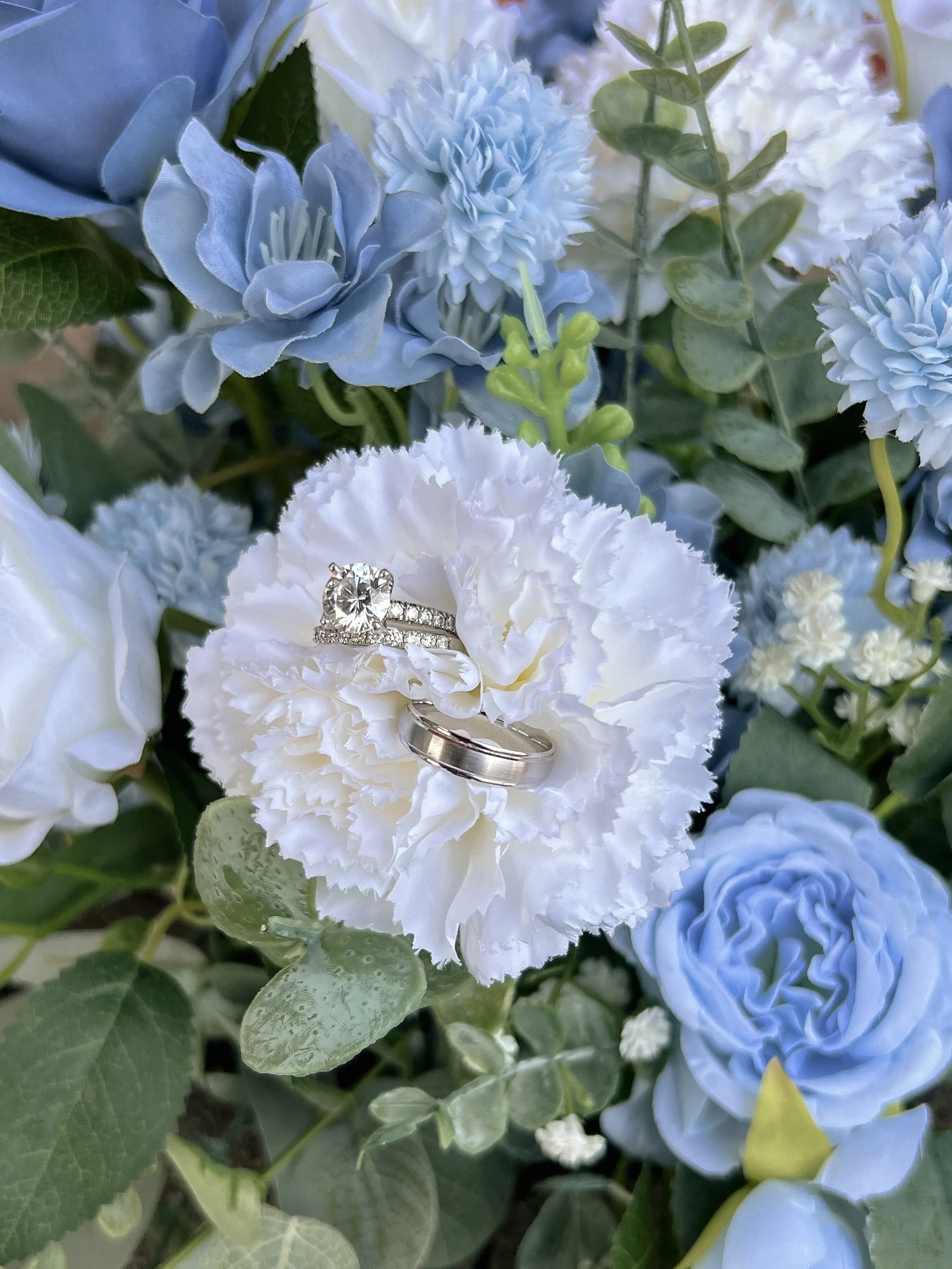 Close-up of blue and white flowers with a diamond engagement ring placed on a white carnation flower.