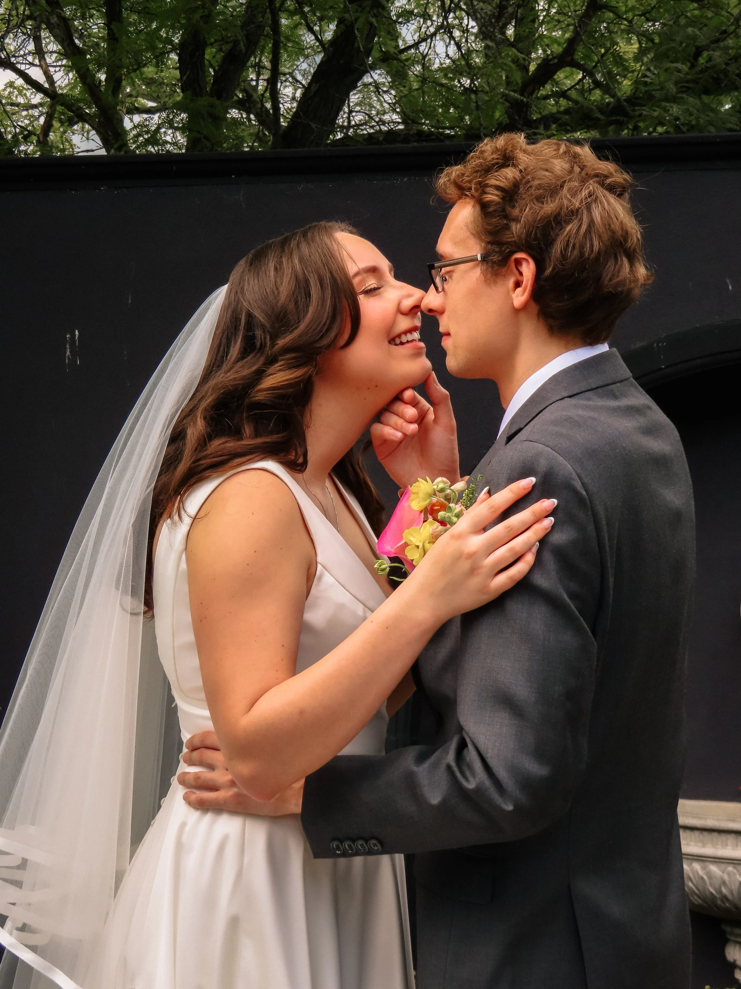 A bride and groom are close together, touching noses and smiling at each other, at their wedding. The bride is wearing a white dress with a veil, and the groom is wearing a gray suit and glasses. The bride is holding a small colorful bouquet, and they appear in front of a dark backdrop with trees overhead.