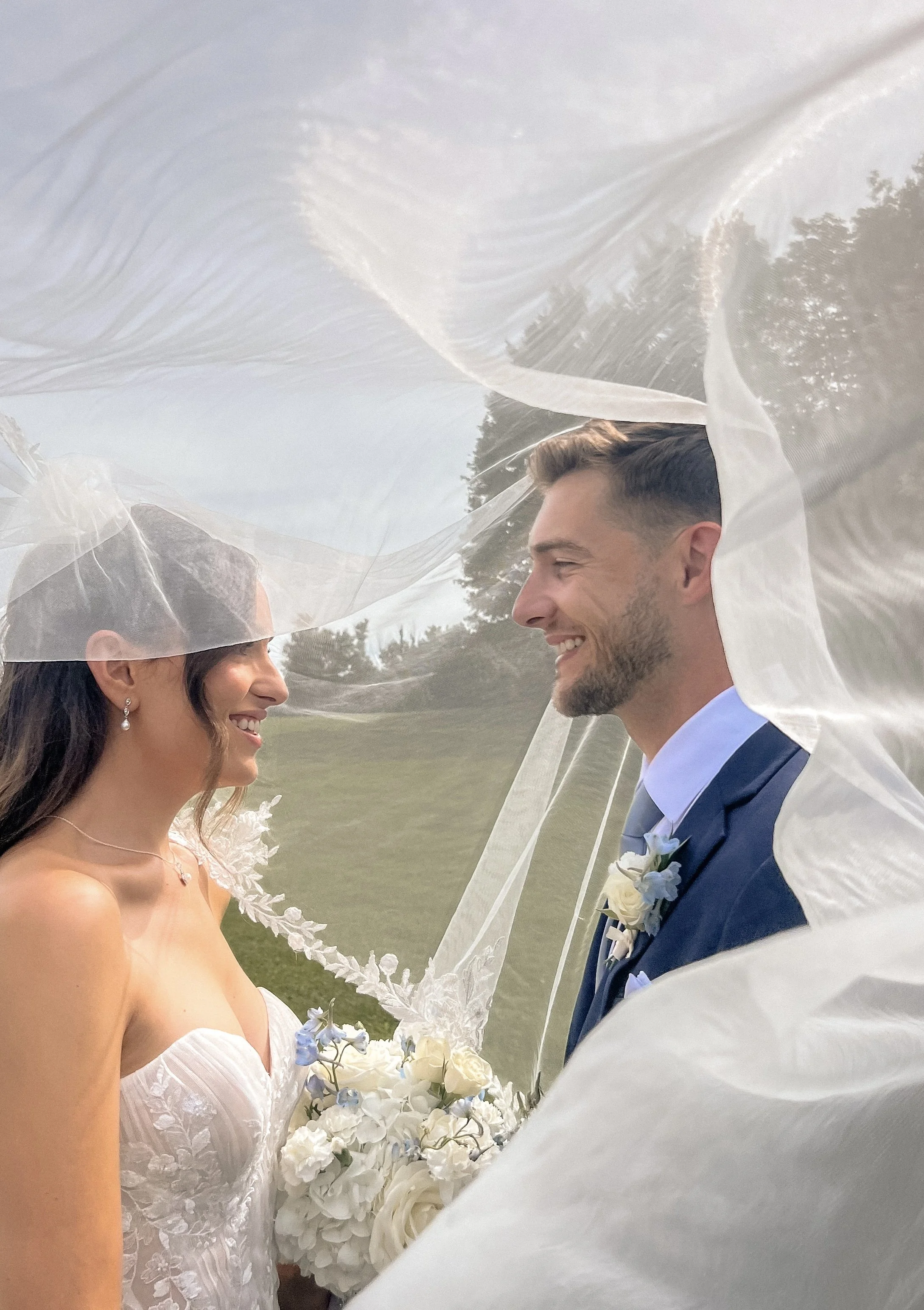 A bride and groom smiling at each other under a sheer veil during their outdoor wedding. The bride is holding a bouquet of white flowers, and they are facing each other with greenery and trees in the background.