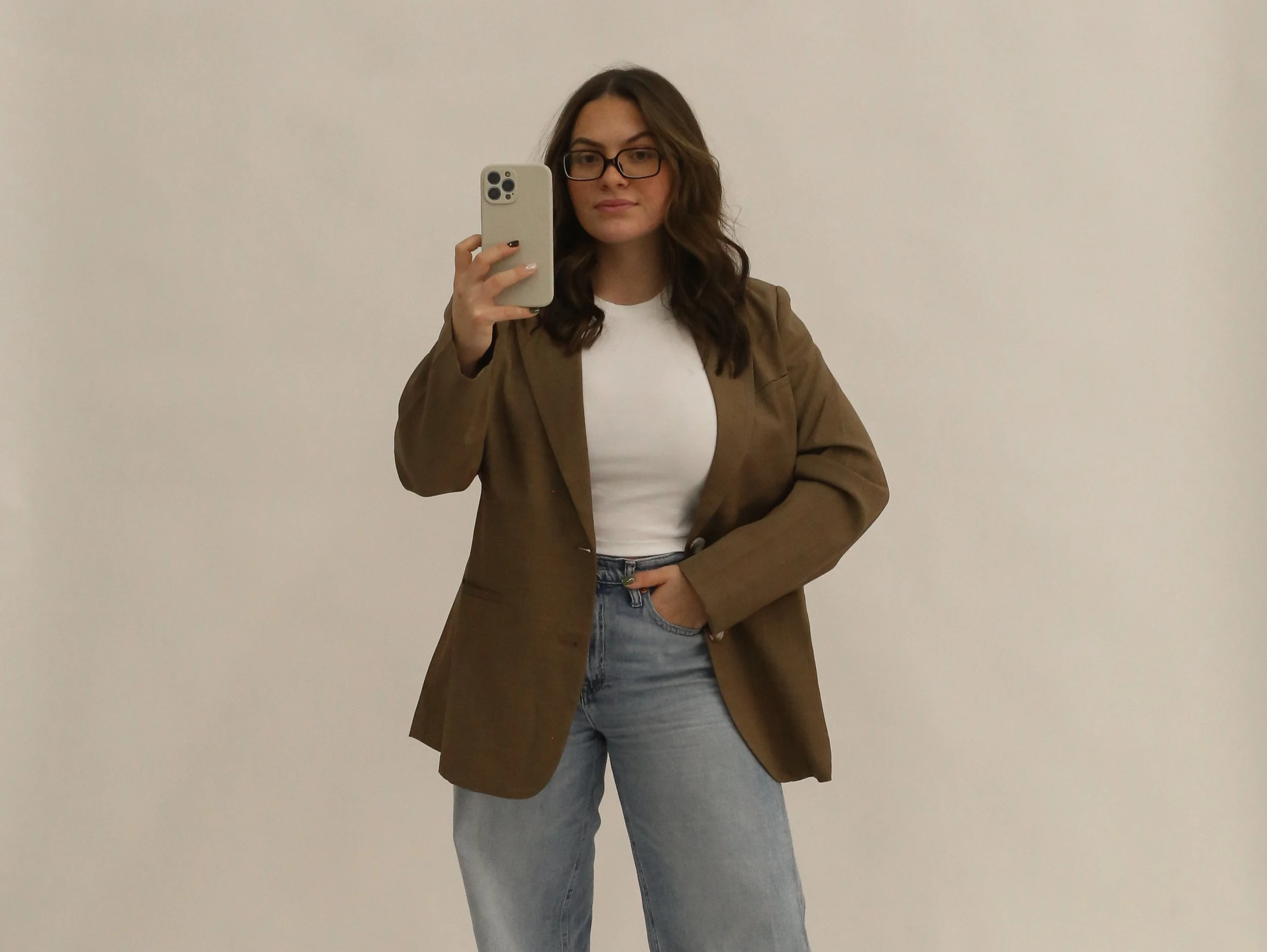 A woman taking a mirror selfie with her smartphone. She has long wavy brown hair, wears glasses, a white shirt, a tan blazer, and blue jeans, standing against a plain white wall.