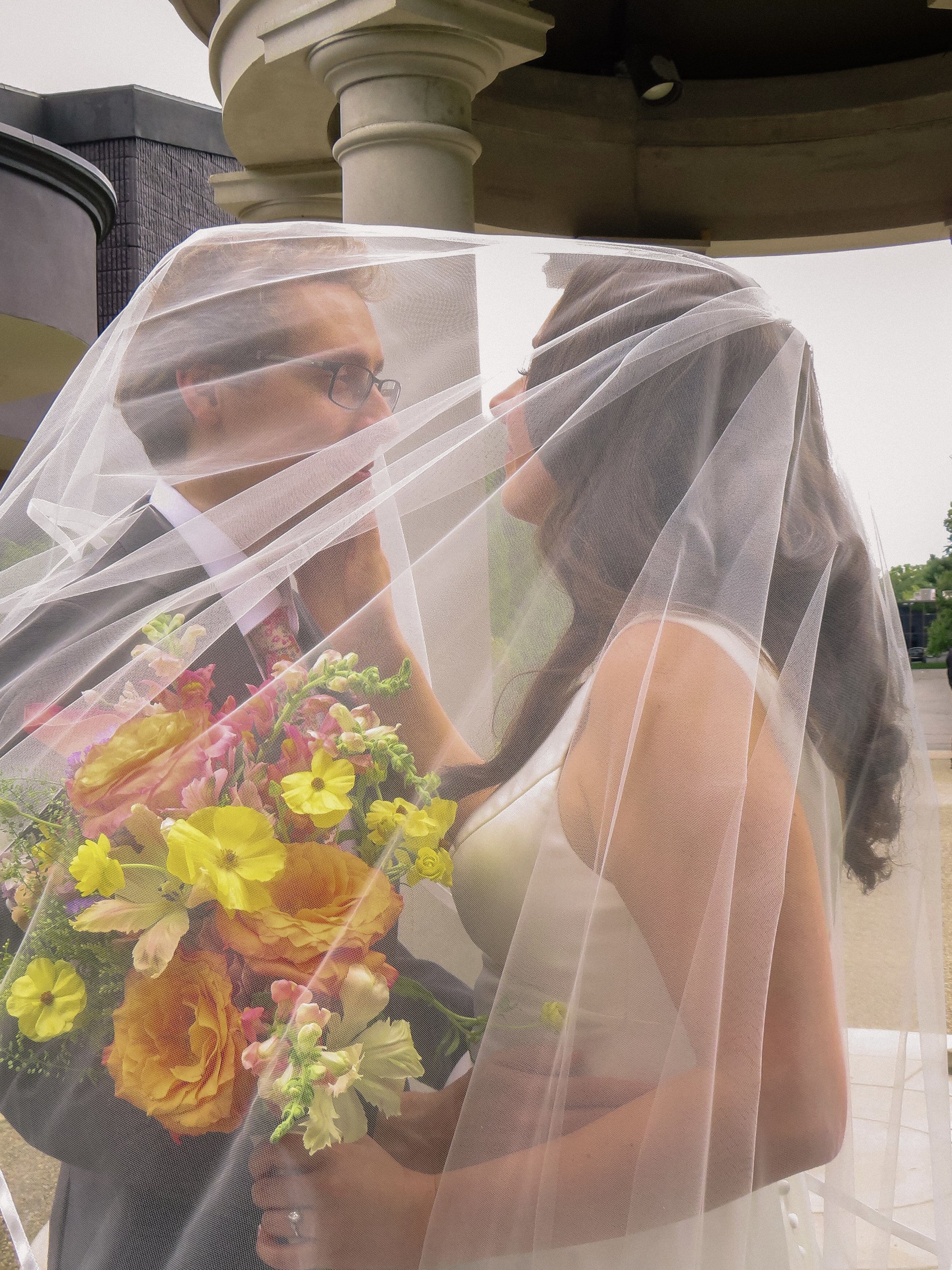 A bride and groom stand face to face, covered by a bridal veil, with the groom holding a bouquet of yellow and pink flowers, outdoors near a building with columns.