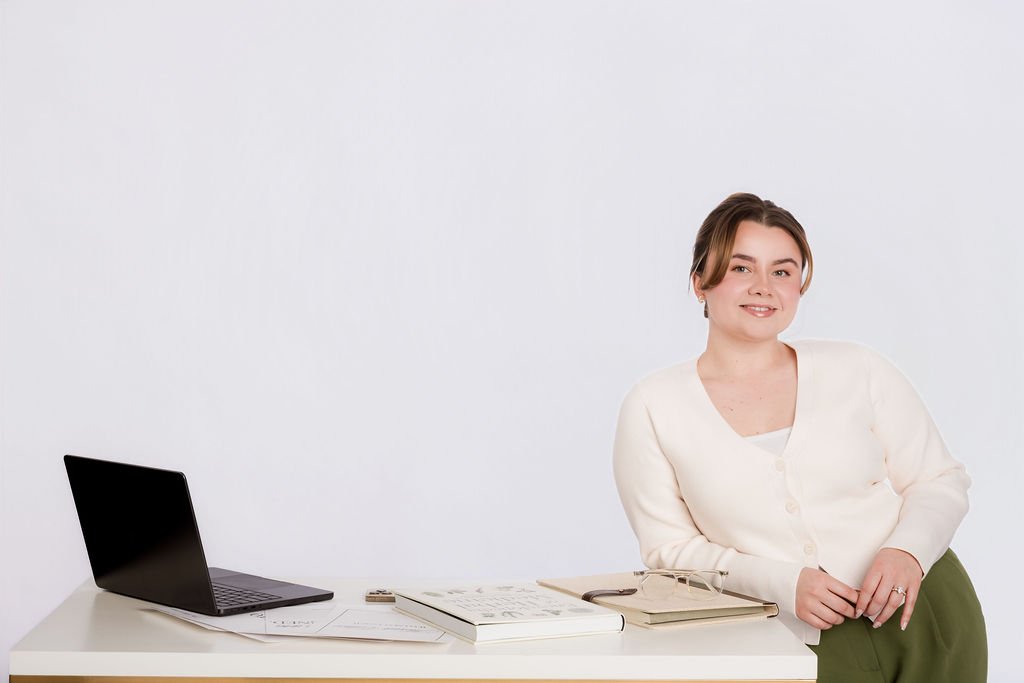 A woman sitting at a desk with a laptop, notebooks, and glasses, smiling at the camera.