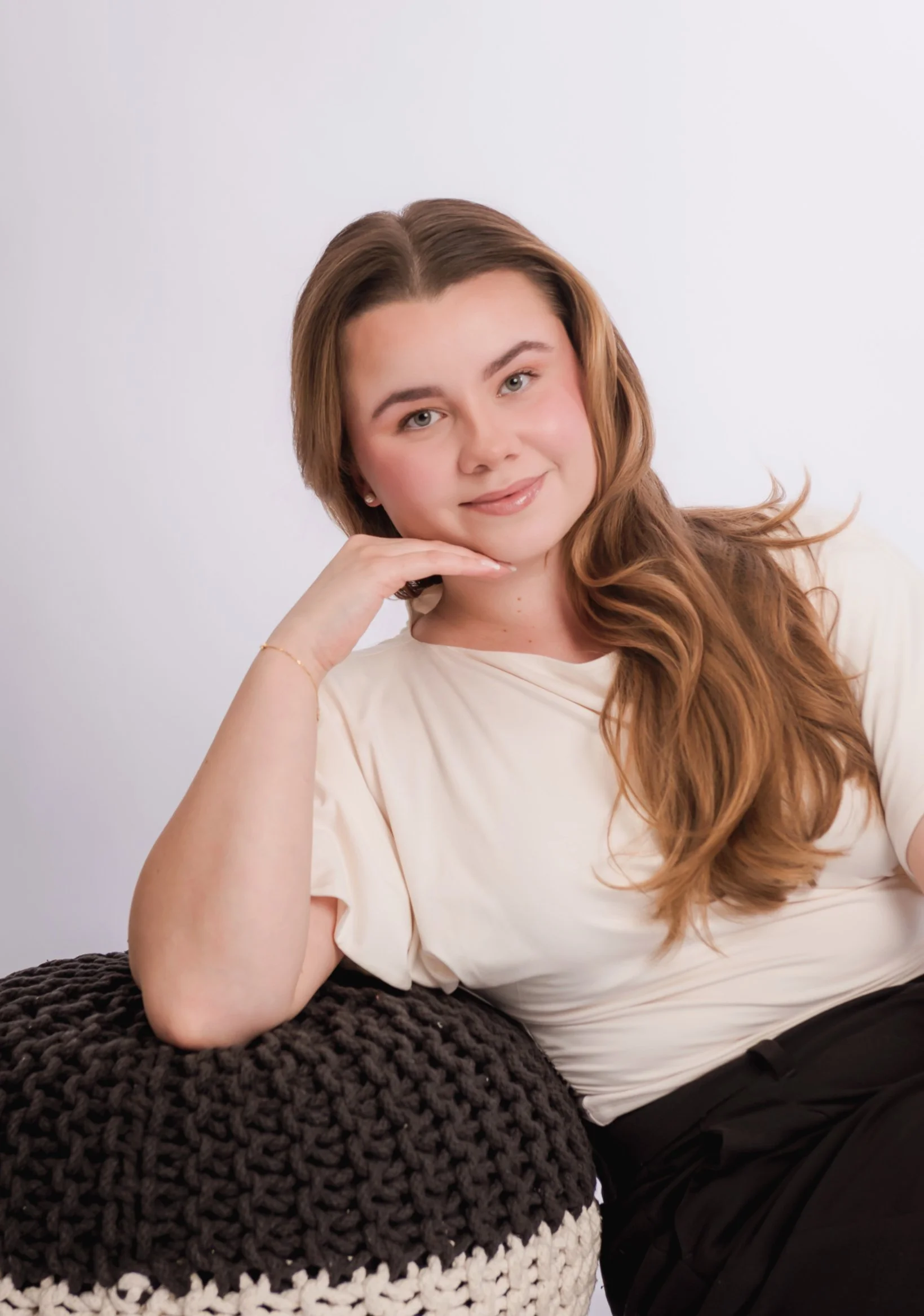 Portrait of a young woman with long, wavy brown hair, smiling, resting her chin on her hand, wearing a white top, leaning on a black textured knitted surface against a plain light background.