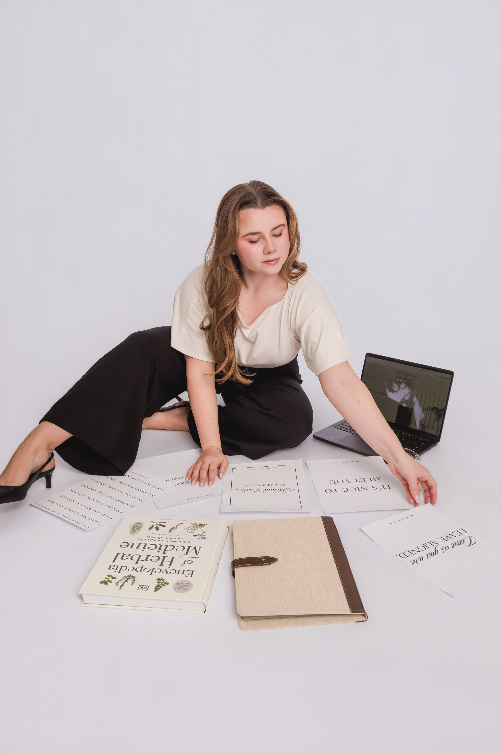 A woman with long wavy hair, dressed in a cream top and black pants, is kneeling on the floor surrounded by papers, books, and a laptop, focusing on organizing or reviewing the materials in a bright, uncluttered space.