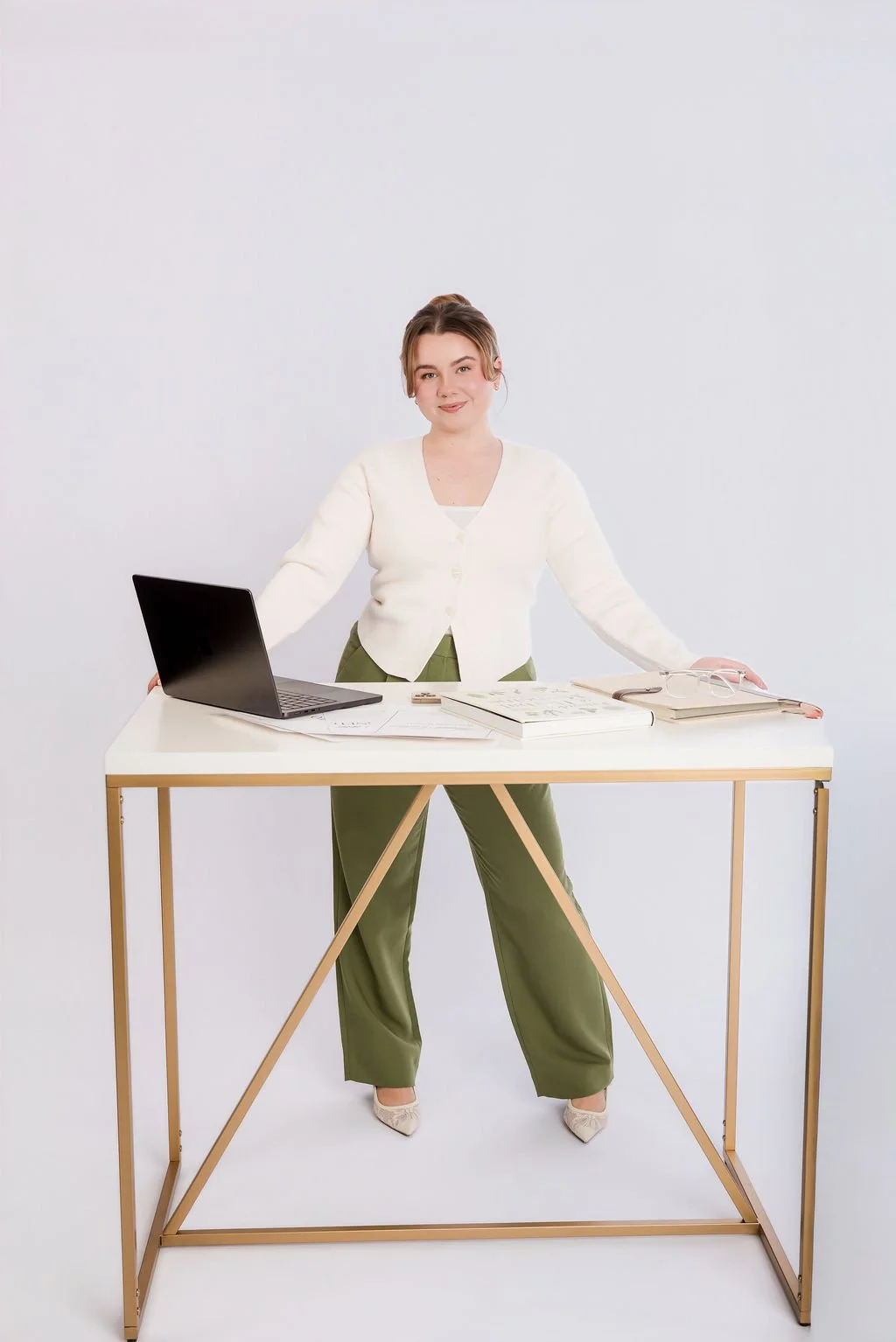 A woman standing behind a white desk with wooden legs, smiling at the camera, with a laptop, closed books, and glasses on the desk, in a plain white background.