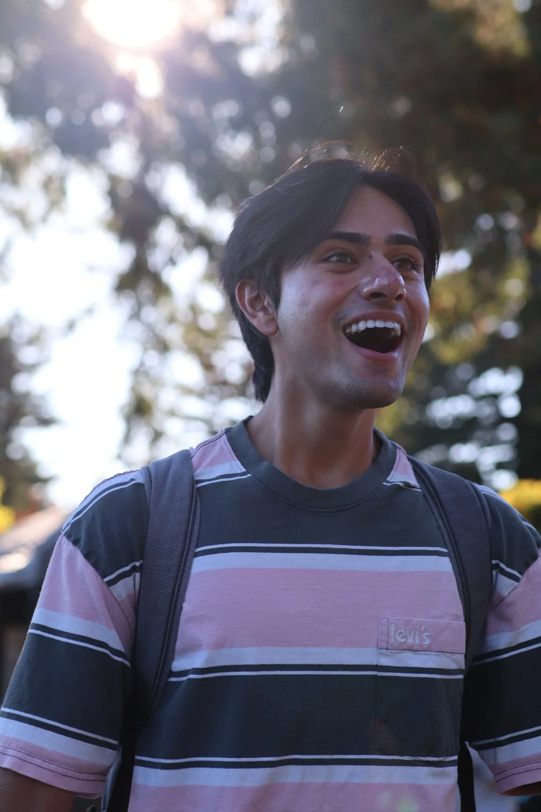 A young man with dark hair smiling outdoors, wearing a striped Levis shirt and a backpack, with sunlight and trees in the background.