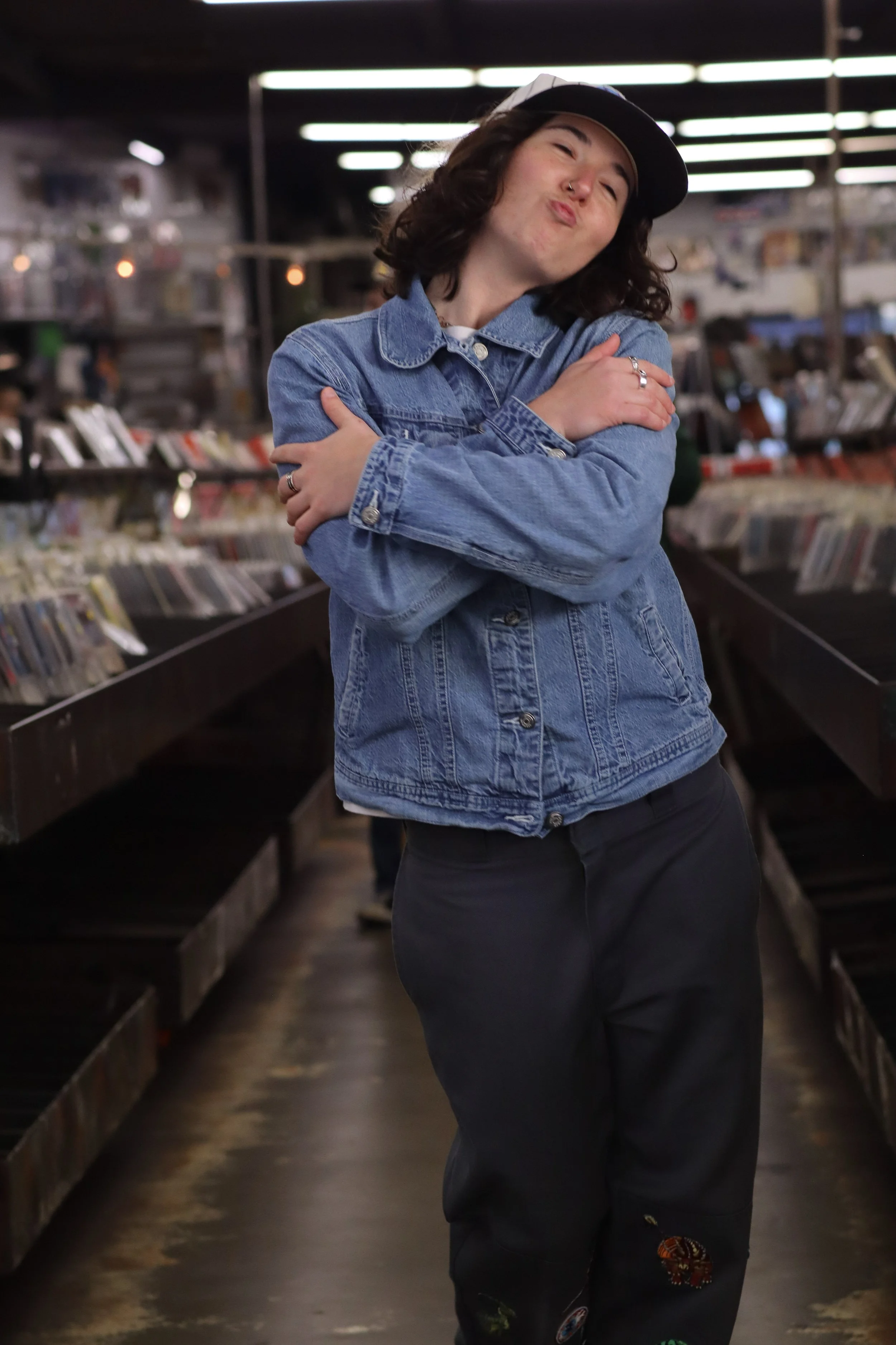 Young woman with curly hair wearing a denim jacket and black cap, posing with arms crossed inside record store with shelves of vinyl records.