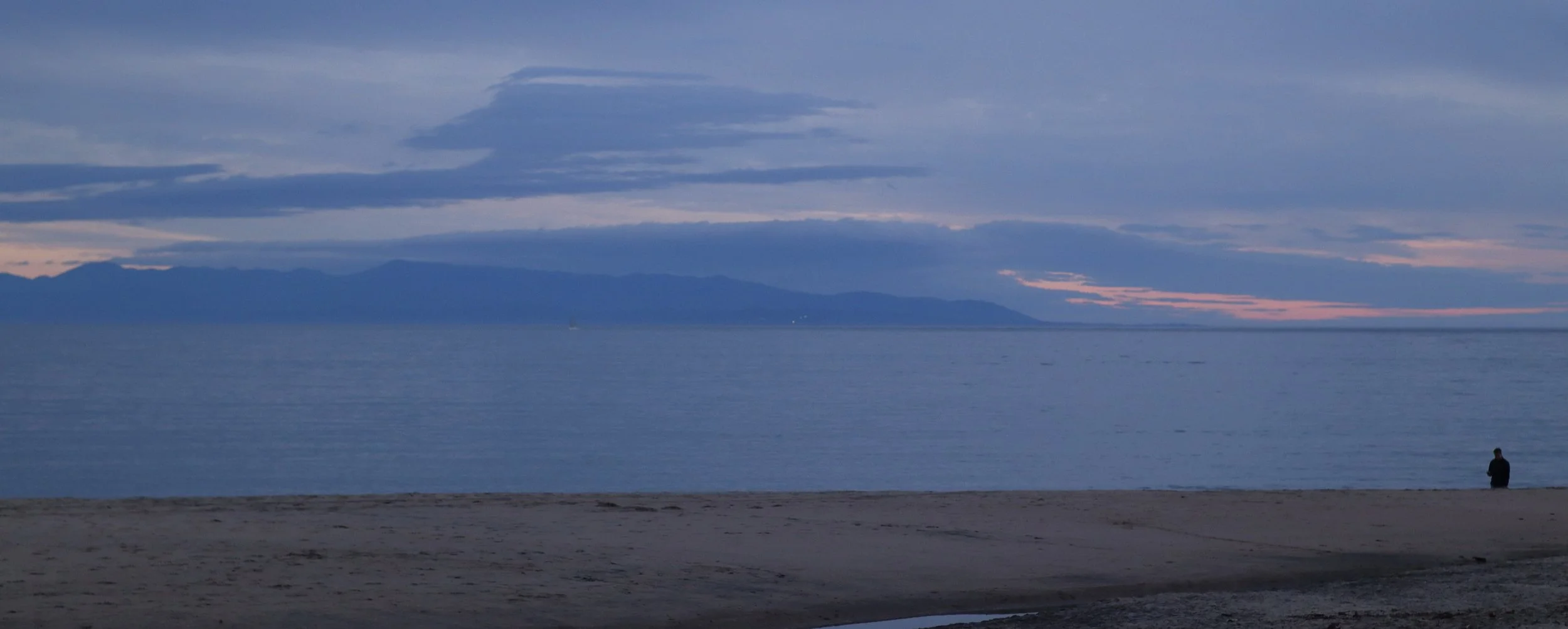 A person sitting on the beach near the ocean at dusk, with mountains and a cloudy sky in the background.