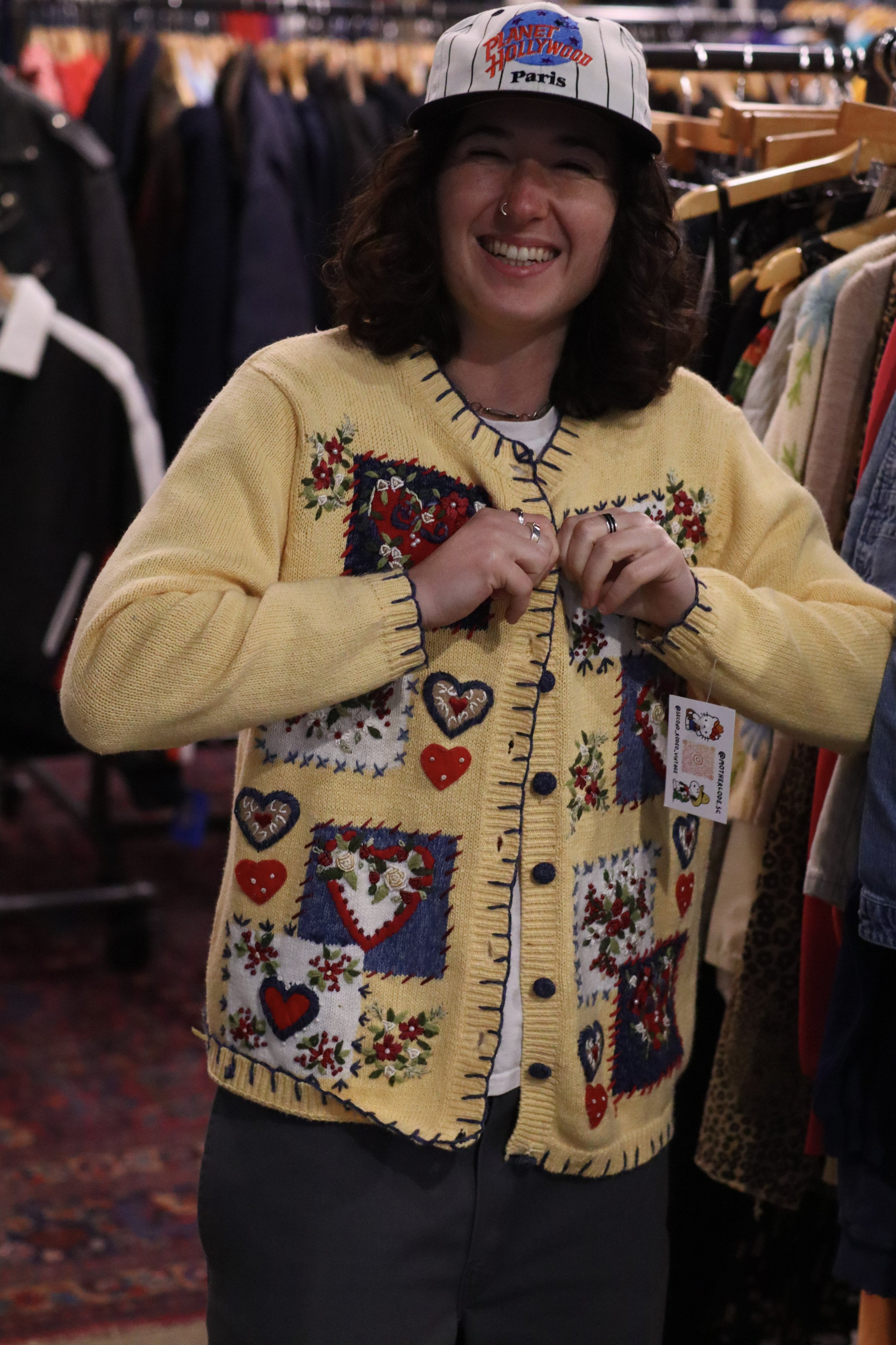 A woman with dark, curly hair and a nose ring, smiling, wearing a yellow embroidered cardigan with hearts and flowers and a white shirt underneath, standing in a clothing store.