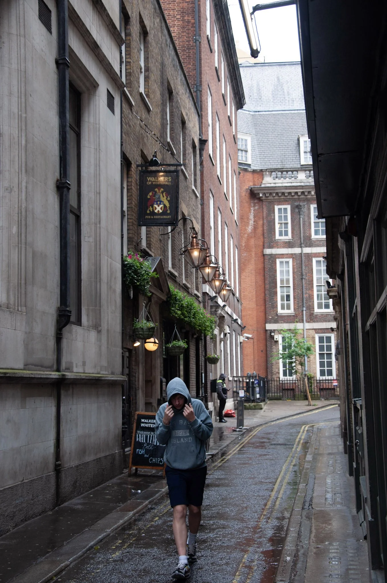 A person walking on a wet cobblestone street in an alleyway, wearing a gray hoodie with the hood up, black shorts, and sneakers. The alley is lined with tall brick and stone buildings, hanging lamps, and potted plants. In the background, there are ot