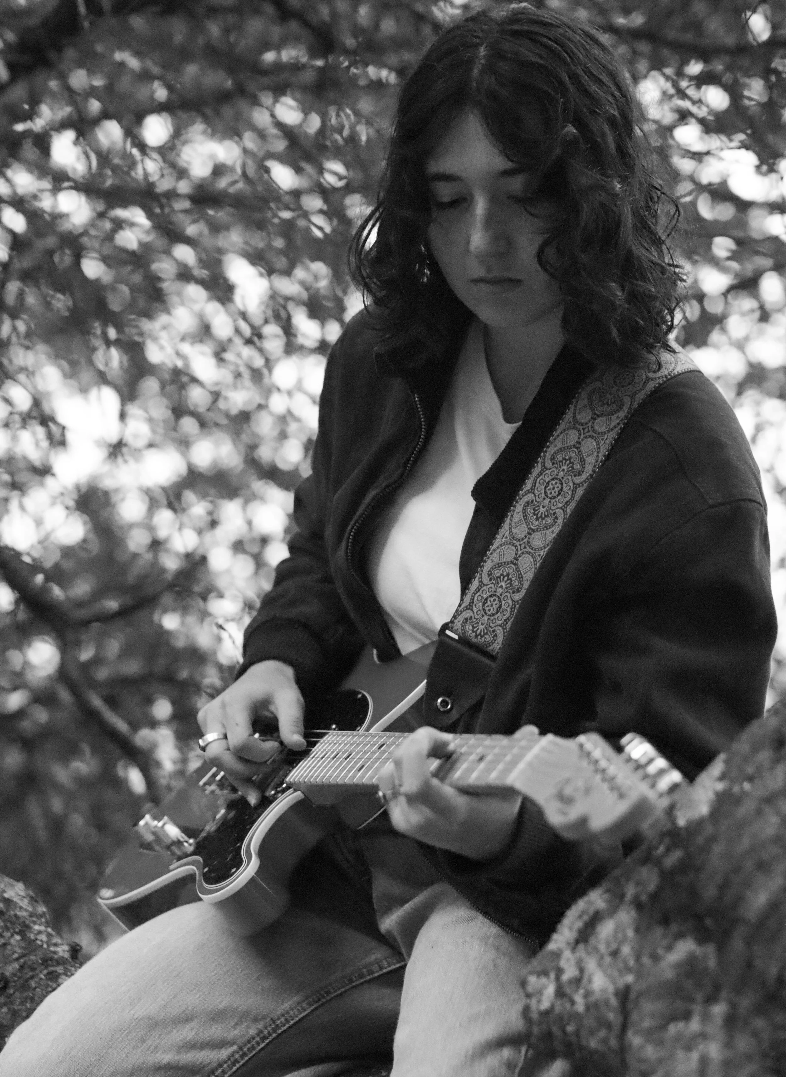 A woman with dark curly hair playing an acoustic guitar outdoors, looking down at the guitar, with leafy background.