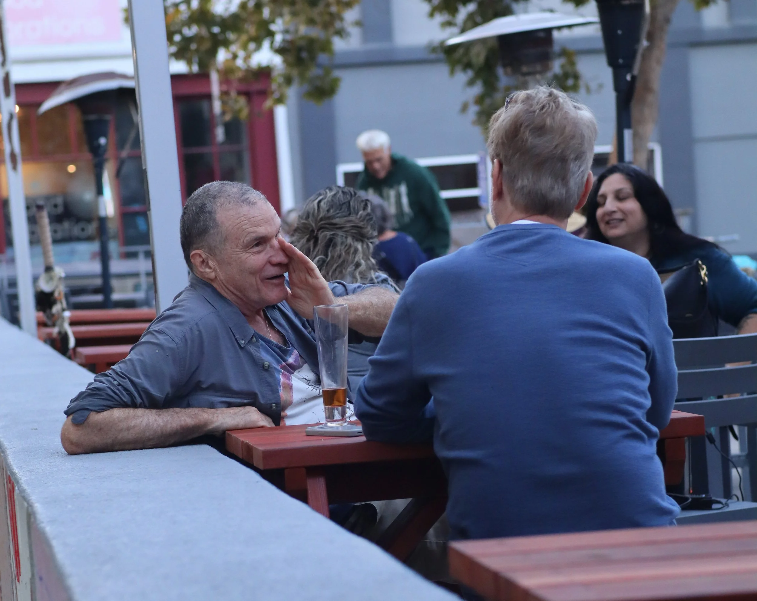 People sitting at outdoor tables, talking and drinking beverages, in a casual gathering setting with trees and buildings in the background.