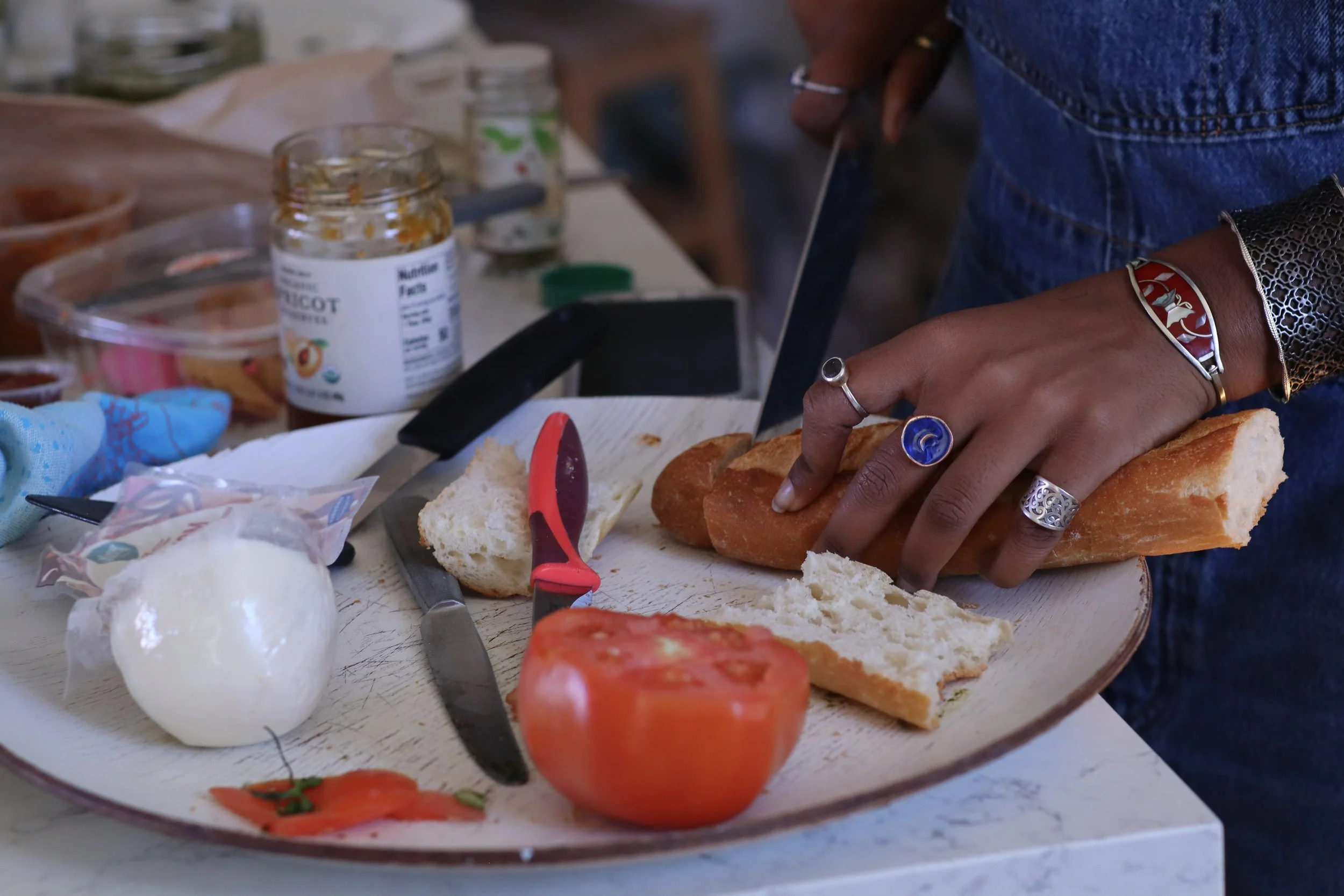 Person slicing bread on a white cutting board, surrounded by tomatoes, mozzarella cheese, and a knife, with jars and a glass of water in the background.