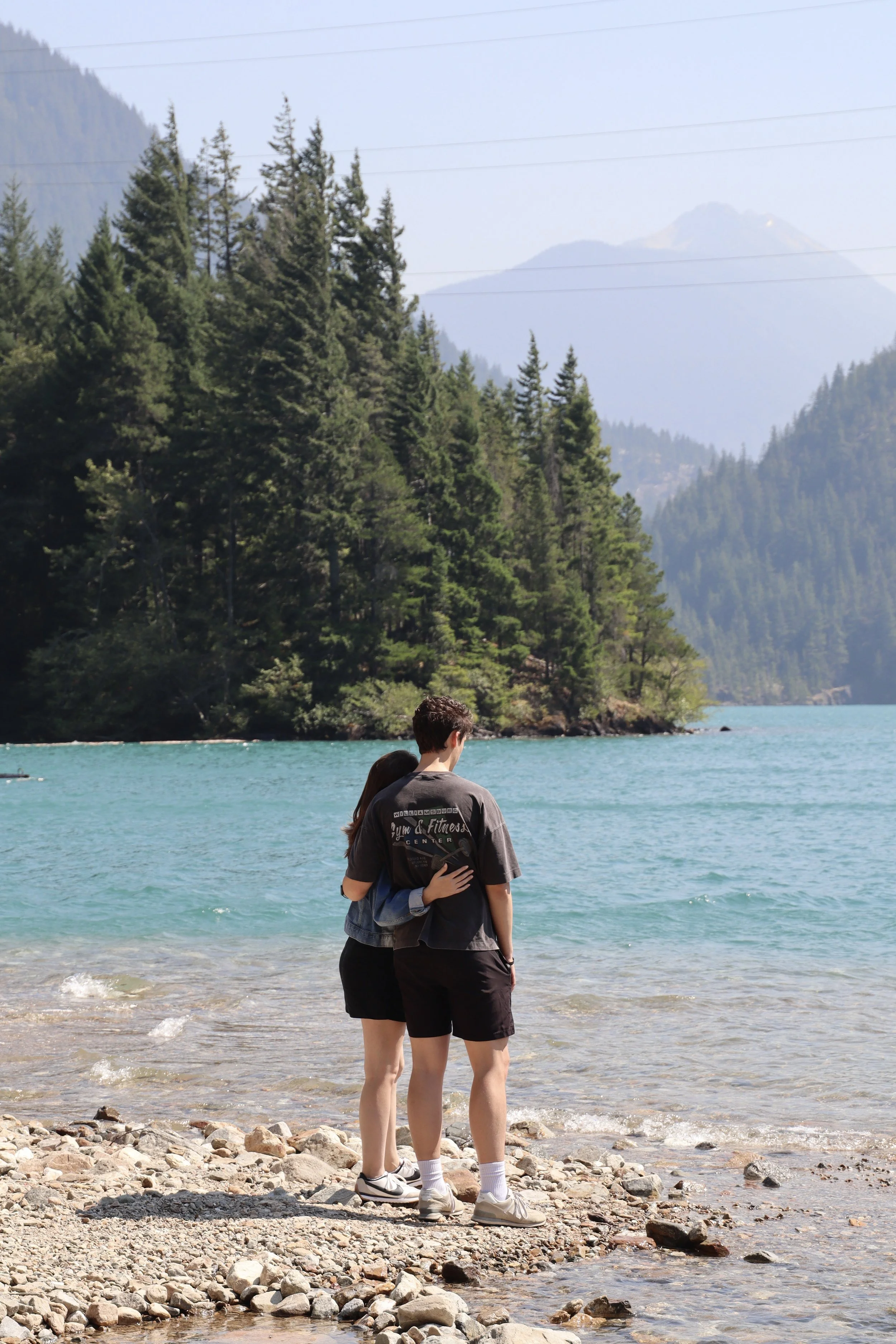 A couple standing together on a rocky lakeshore, embracing each other, with mountains, trees, and a lake in the background.