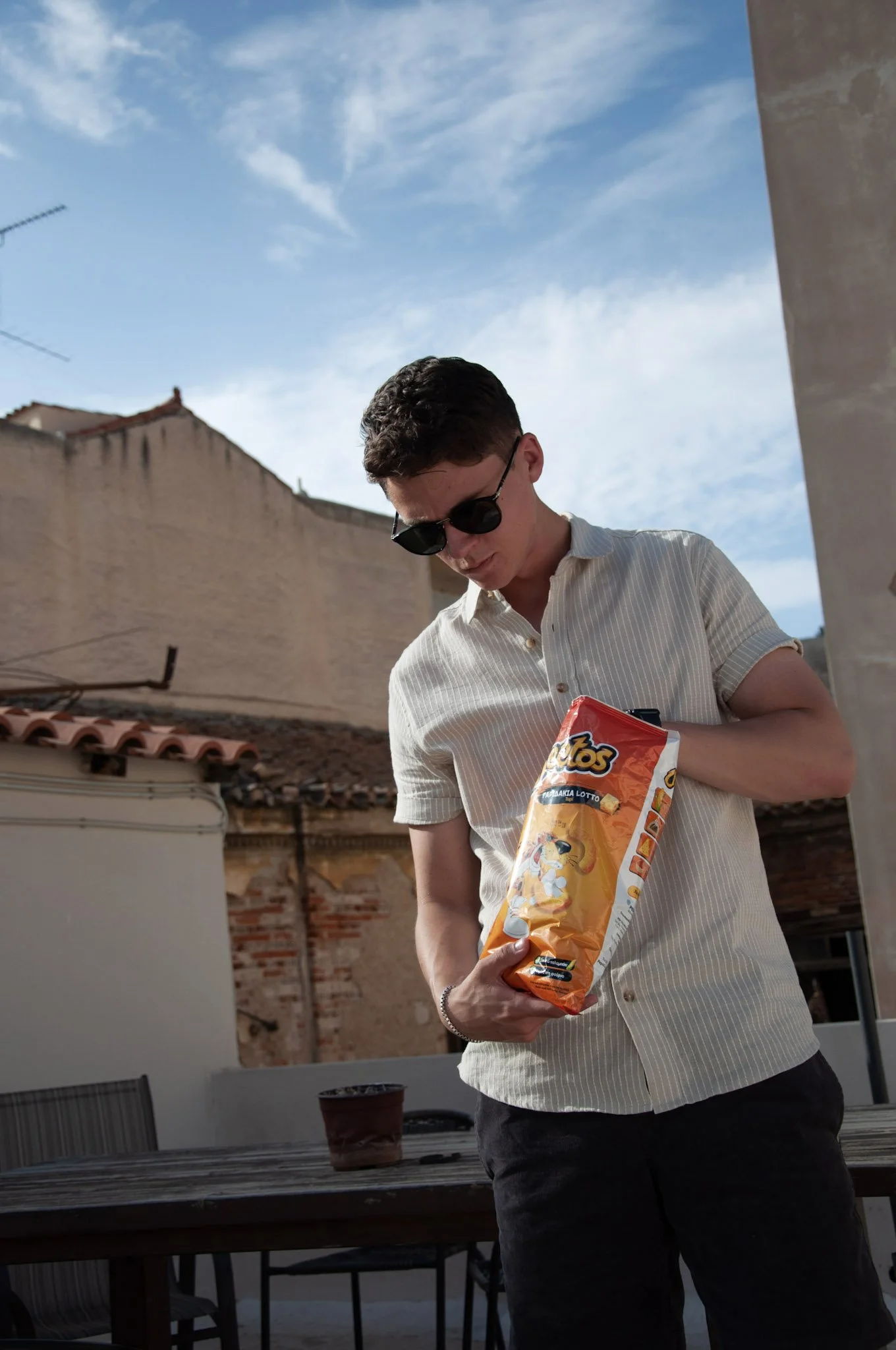 A young man with short dark hair wearing sunglasses, a light striped short-sleeve shirt, and dark pants is standing outside on a terrace or balcony, holding an open bag of potato chips.