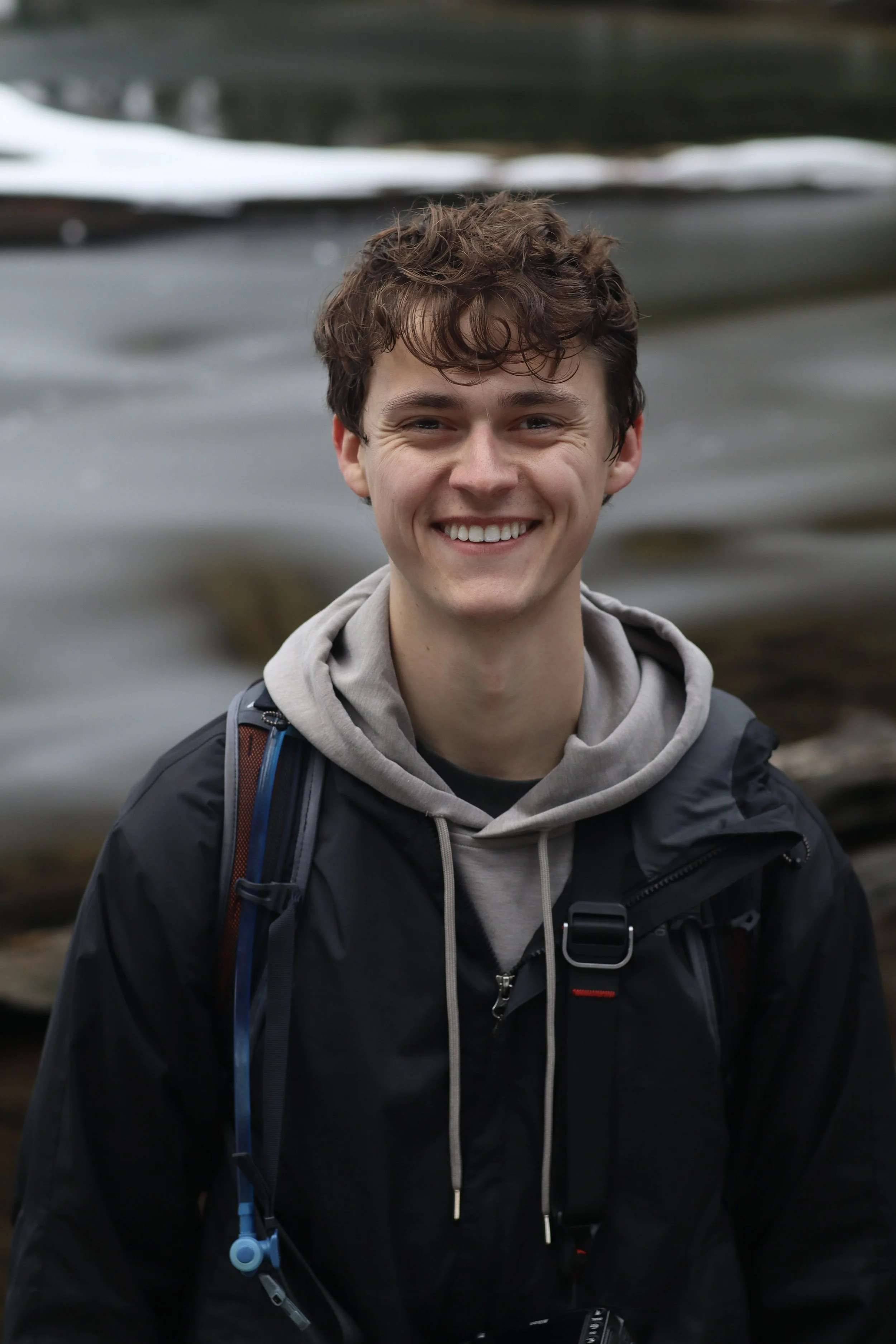 A young man with curly brown hair smiling outdoors near a body of water, wearing a black jacket, gray hoodie, and carrying a backpack.