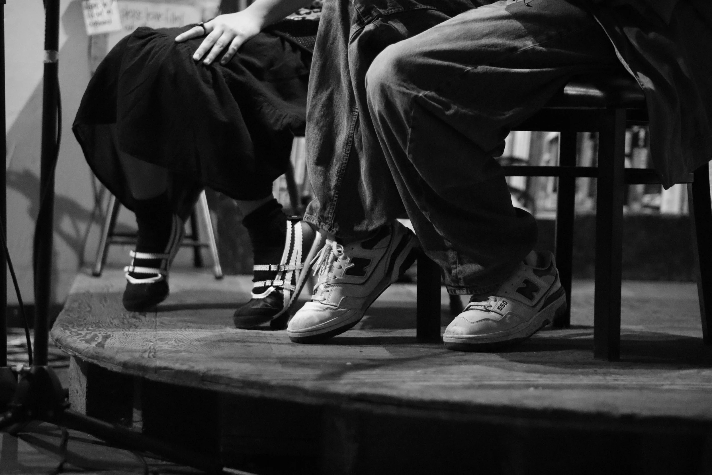 Black and white photo of people sitting on chairs on a stage, with only their lower bodies visible, including legs, feet, and hands resting on laps or chairs.