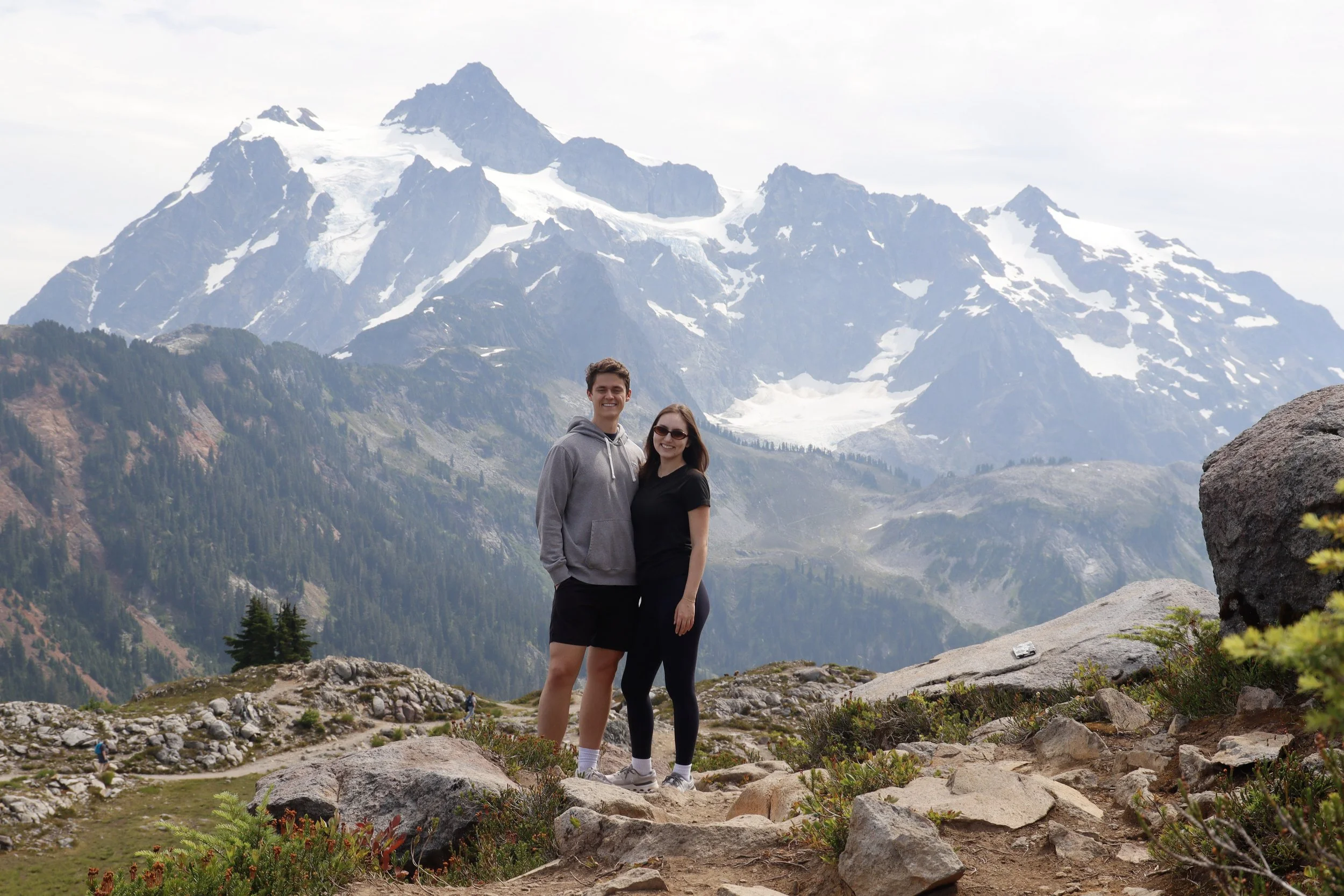 A smiling young man and woman standing together in a mountainous landscape with snow-capped peaks in the background. They are on rocky terrain surrounded by some greenery.