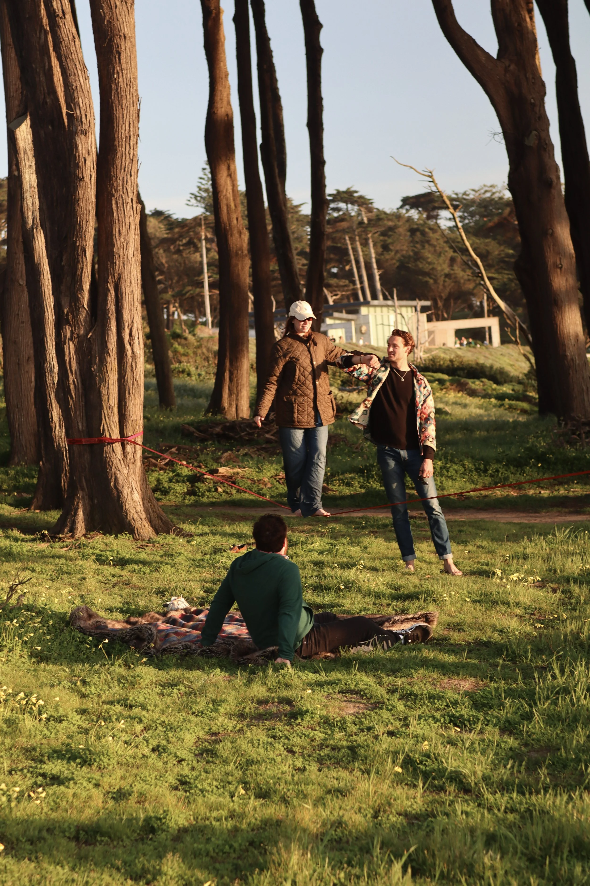 A group of people enjoying a sunset outdoors in a wooded area. Two individuals are walking and holding hands, one in a tan jacket and light hat, and the other in a colorful jacket and black shirt. Another person is sitting on the grass next to a hors