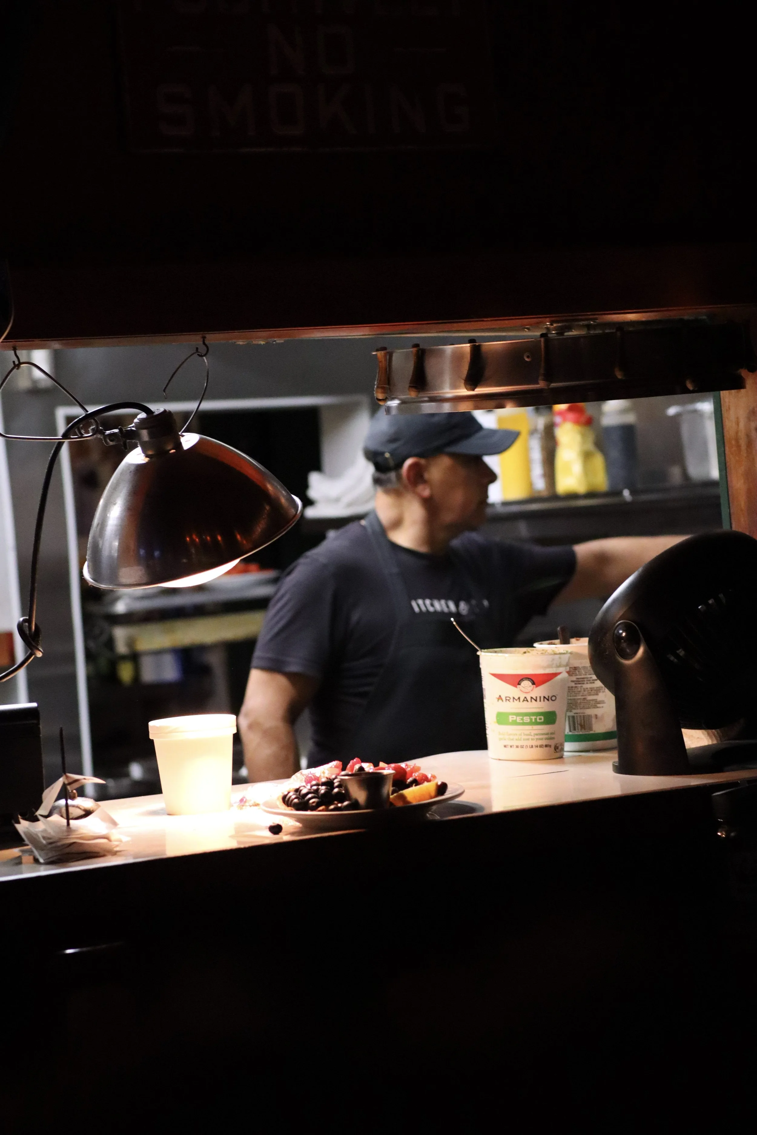 A person wearing a black cap and black t-shirt preparing food in a kitchen with dark shelving, a yellow bottle in the background, and food on a counter with a cup, fruit, and a container of pesto.