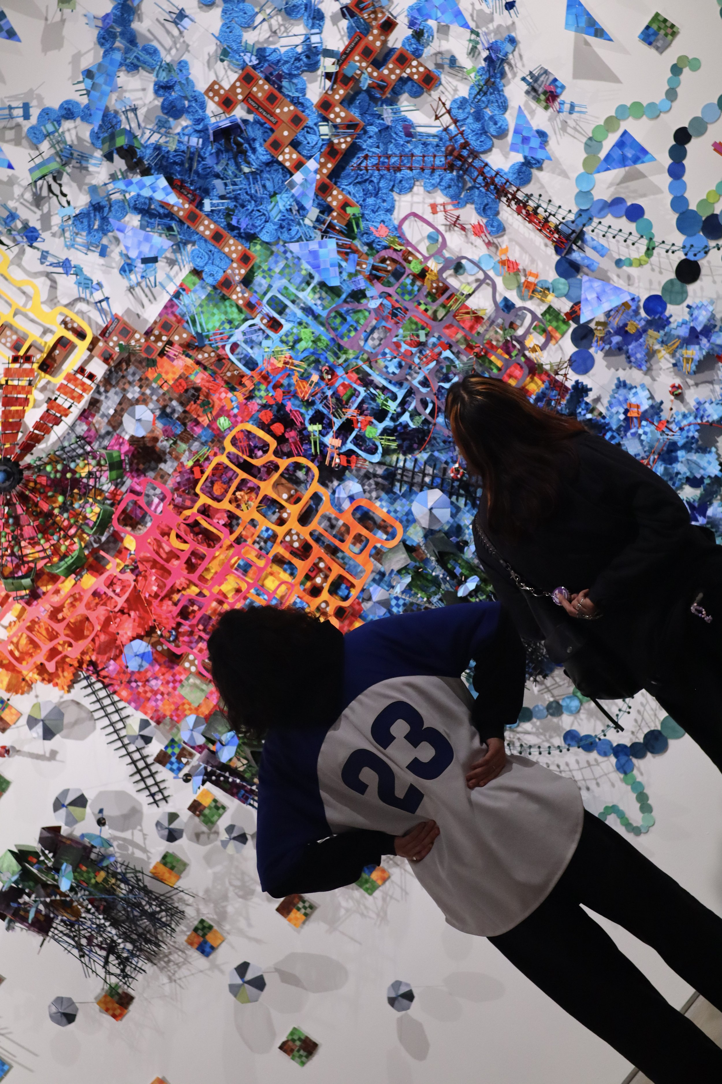 Two people viewing an intricate, colorful, abstract art installation with various geometric shapes, textures, and patterns on a white wall.