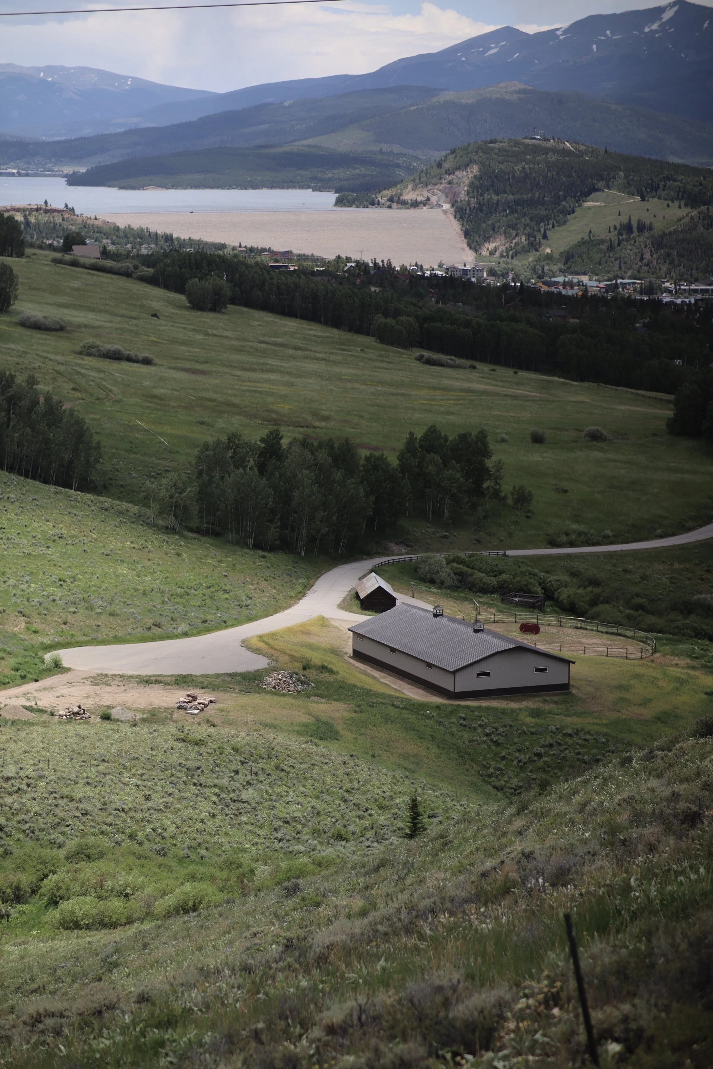 A scenic landscape showing rolling green hills, a small building, and a winding road in the foreground. There are trees scattered across the hills. In the background, a body of water is visible along with forested hills and mountain ranges under a pa