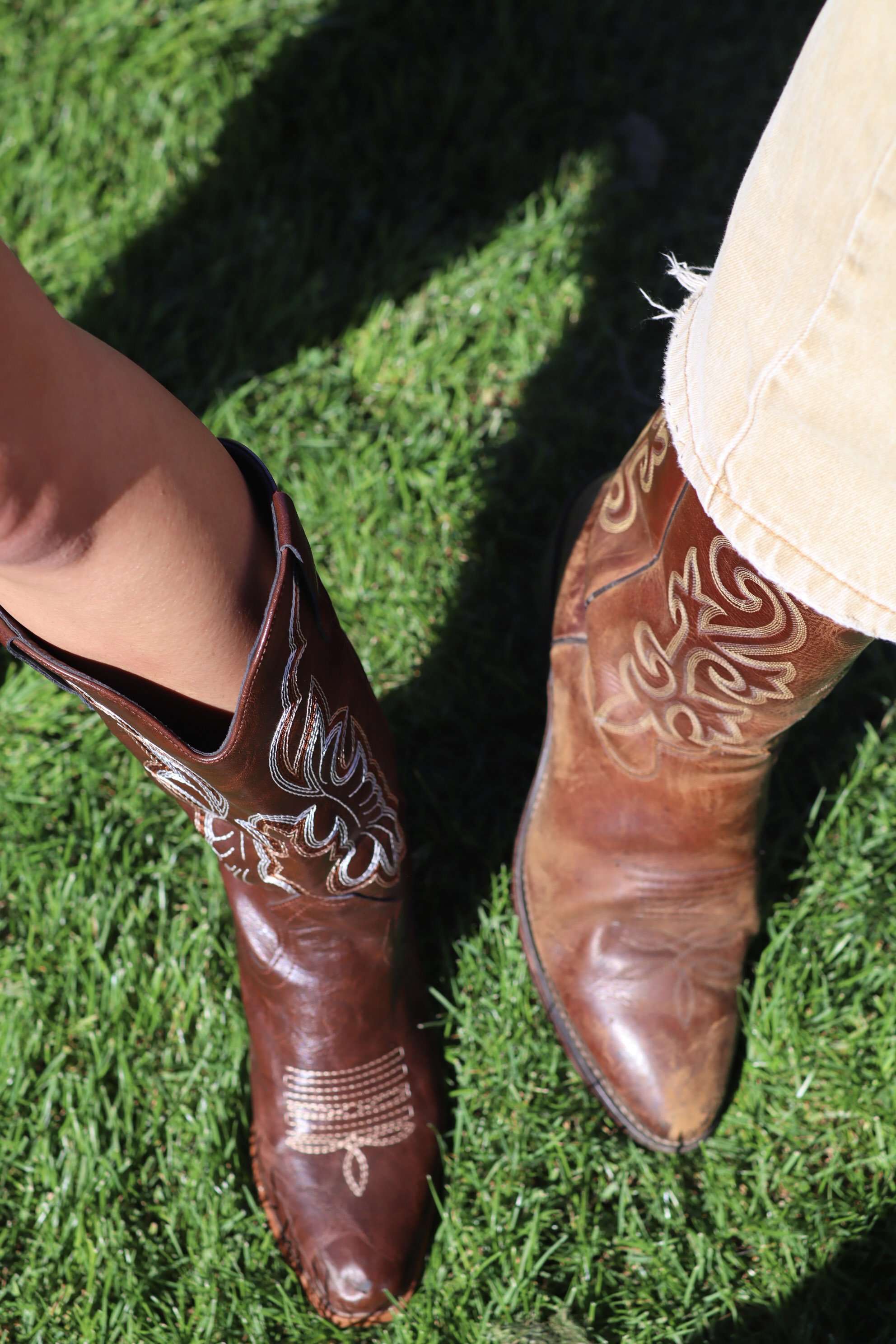 Close-up of two cowboy boots on green grass, one brown with white decorative stitching and the other light tan with embroidered details.