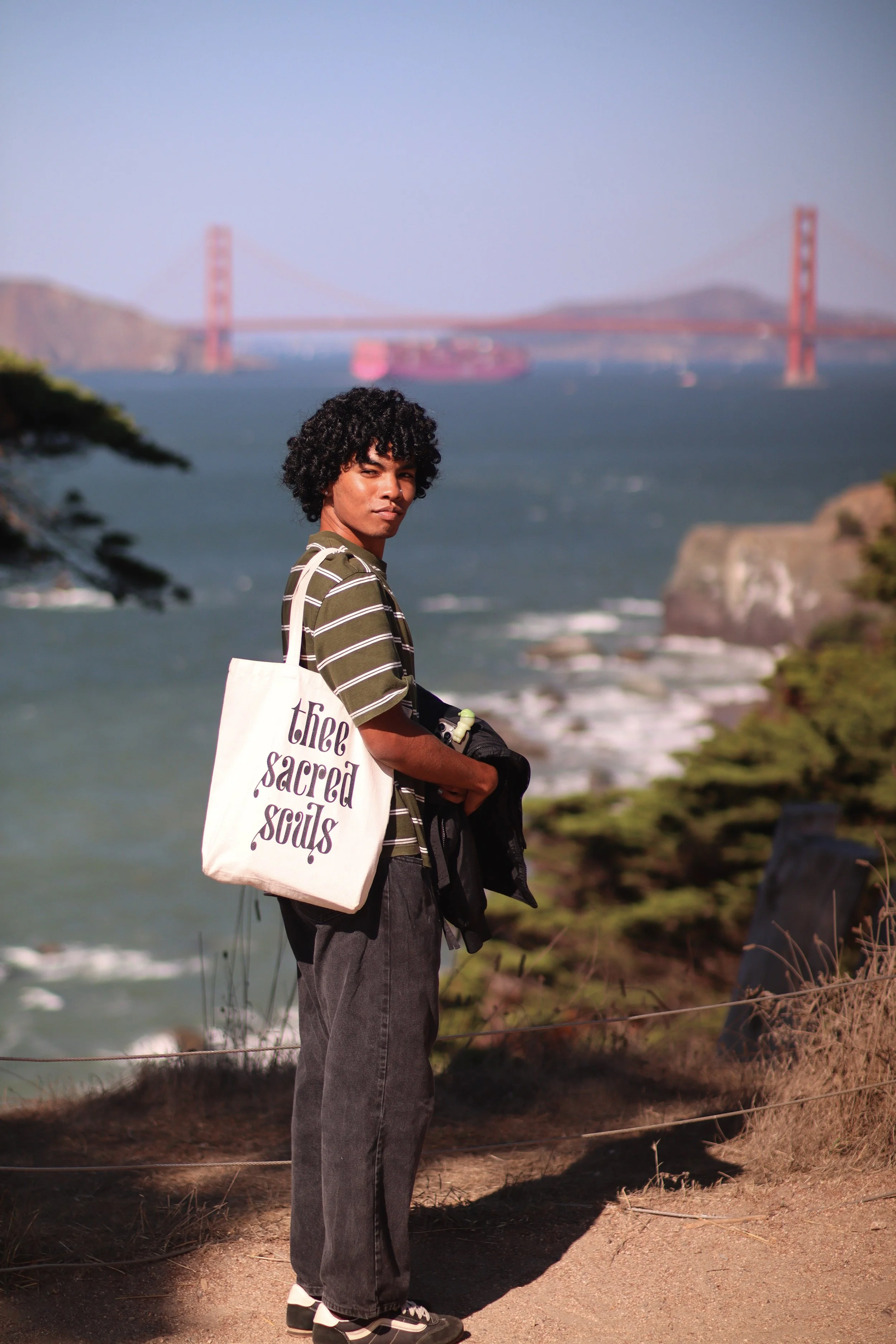 A young man with curly hair standing near a fence by the coast, with the Golden Gate Bridge in the background. He is wearing a striped shirt, dark pants, and sneakers, carrying a tote bag that reads 'the sacred sands.'