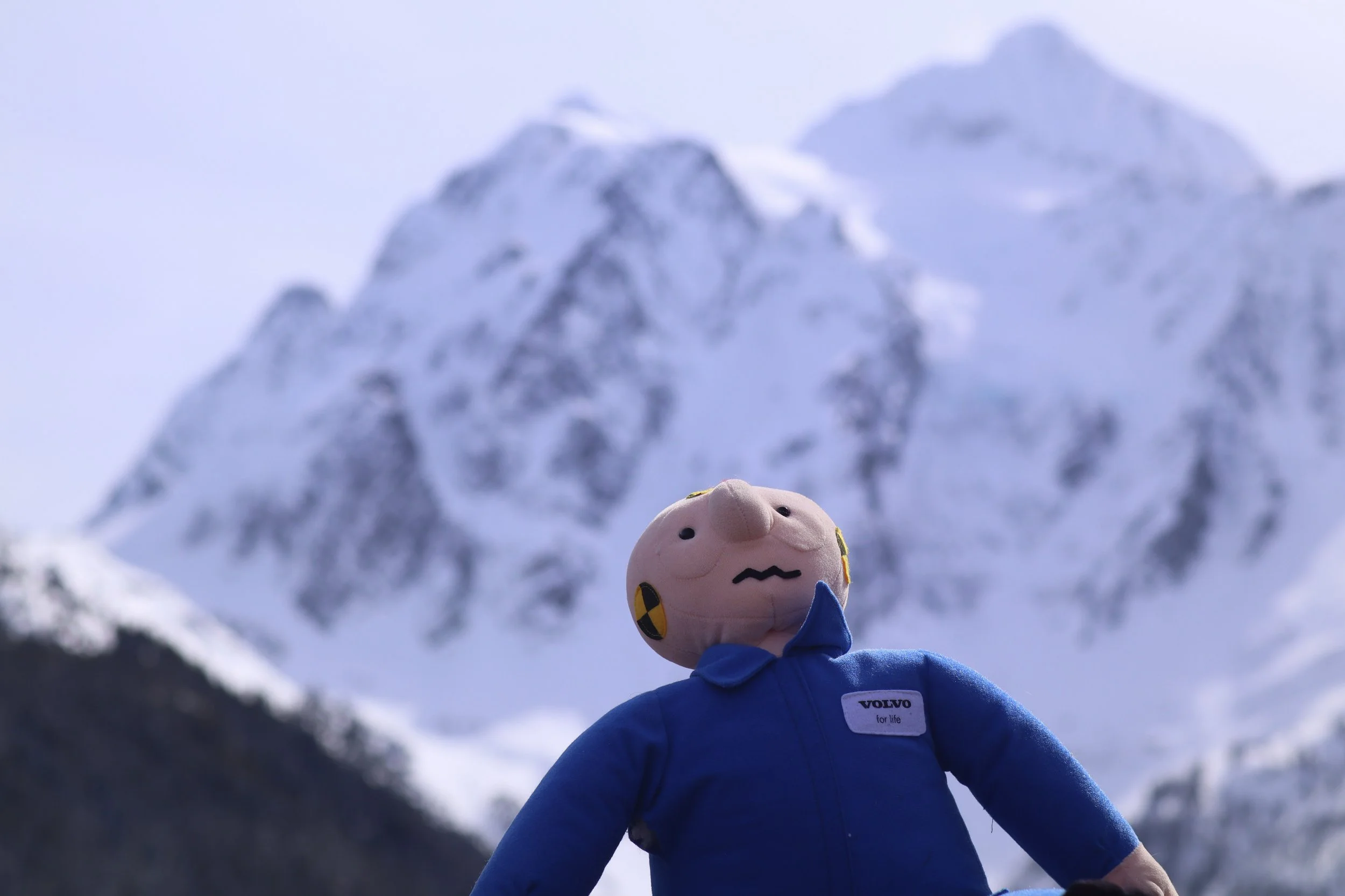 A plush toy character wearing a blue outfit and a Volvo tag, standing outdoors in front of snow-covered mountains with cloudy skies.